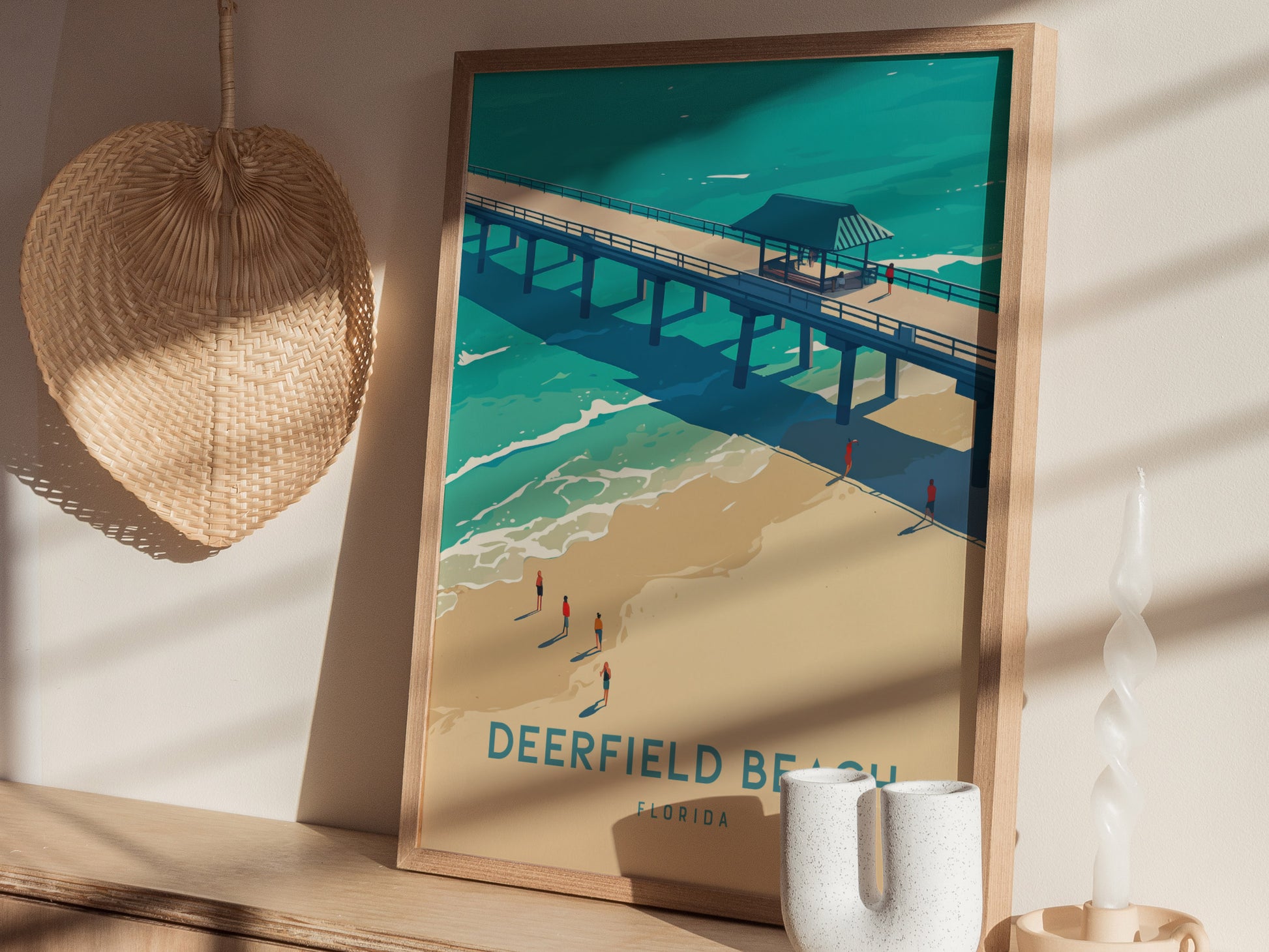 A framed Deerfield Beach poster leans against a wall, depicting a pier over turquoise waters and golden sands. Nearby, a woven hat.