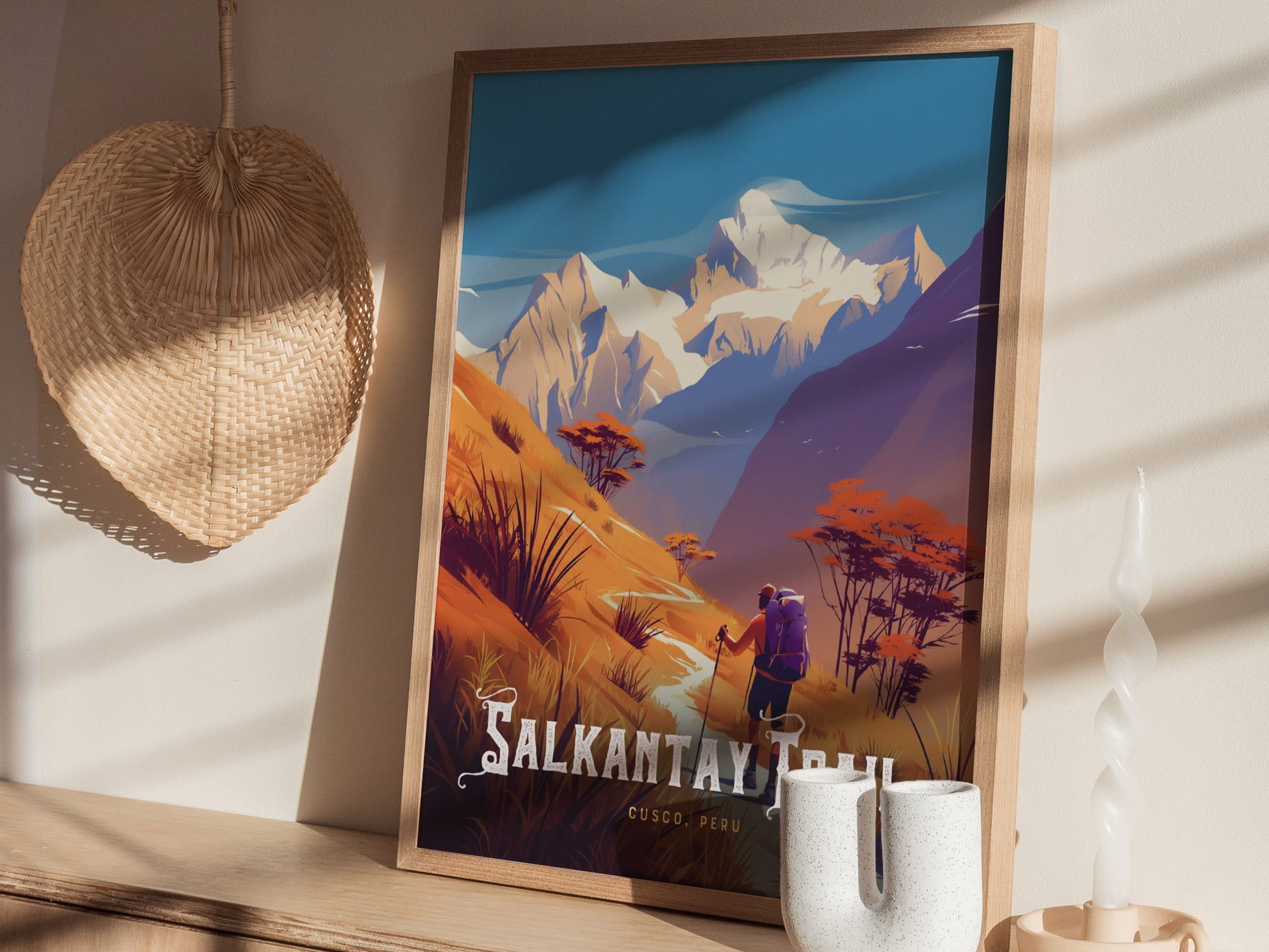 A framed poster of the Salkantay Trail features vibrant illustrations of hikers amidst Andean peaks. It rests on a wooden surface alongside a woven wall decoration and decorative candle.