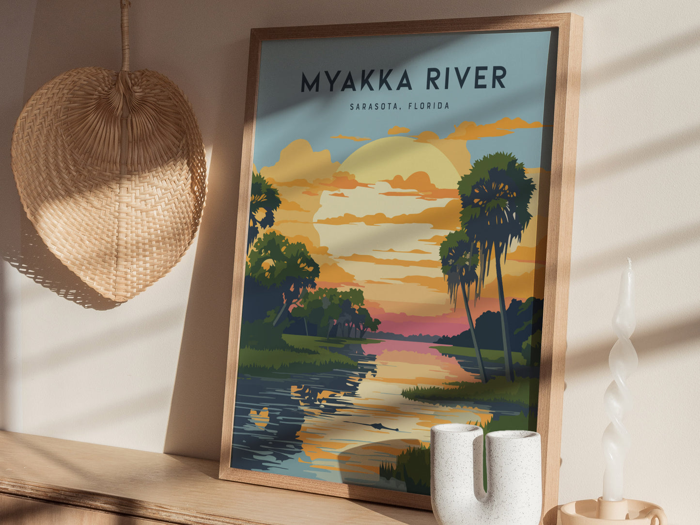 A framed poster of Myakka River, Sarasota, Florida, features a colorful sunset with palm trees and a tranquil river. Its placed on a wooden shelf beside a wicker decoration and.