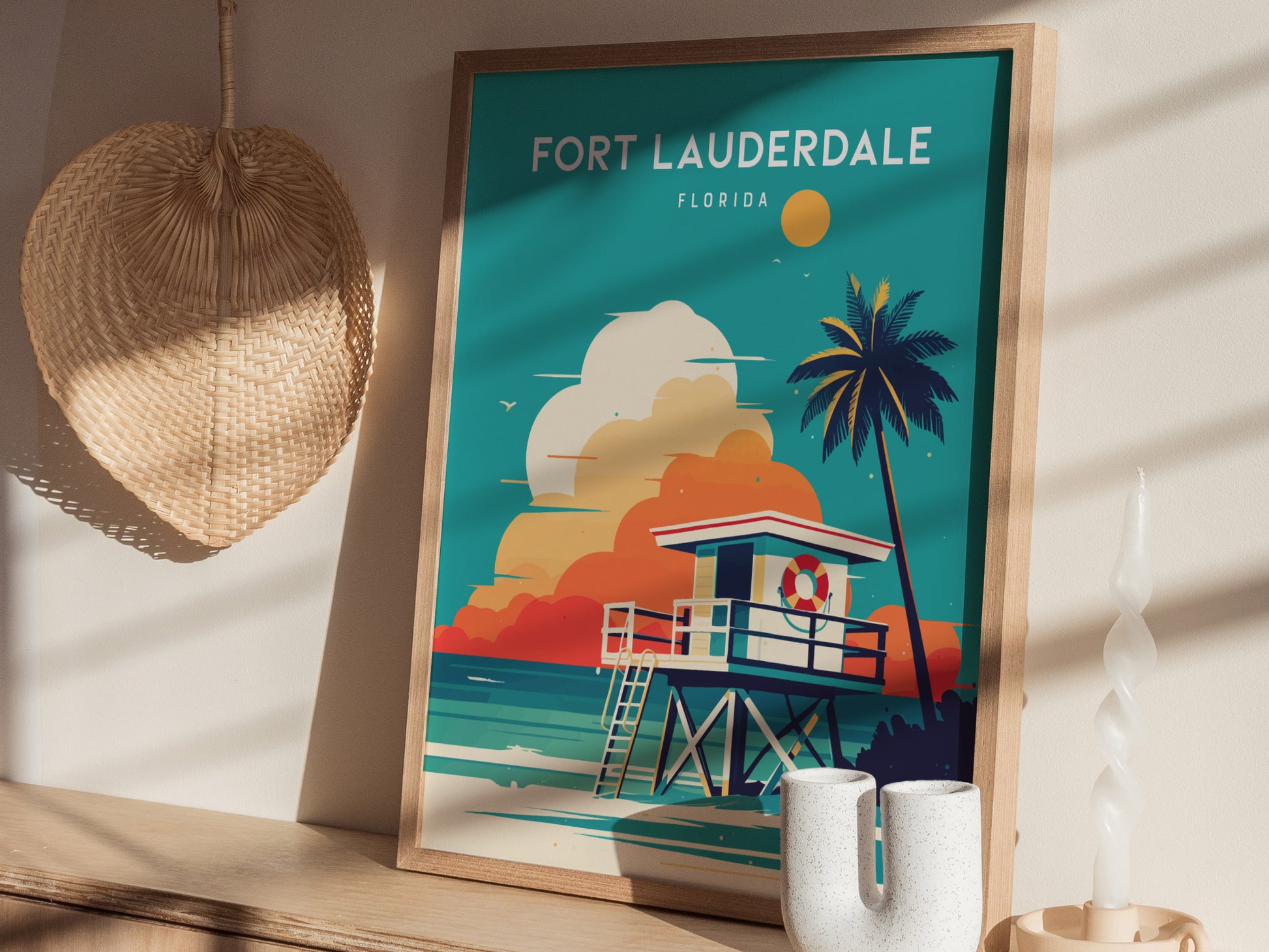 A framed Fort Lauderdale poster with a coastal scene, featuring a lifeguard stand and palm tree, is displayed on a shelf beside a woven wall decor and a twisted candle.