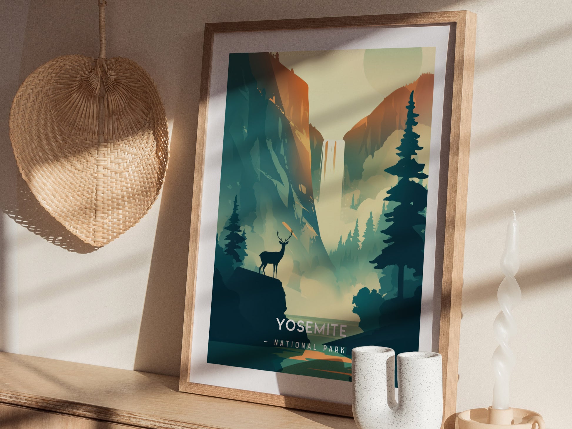 A framed Yosemite National Park poster with a minimalist design is displayed on a wooden shelf, featuring a stag silhouette and pine trees. A woven fan and candle are nearby.