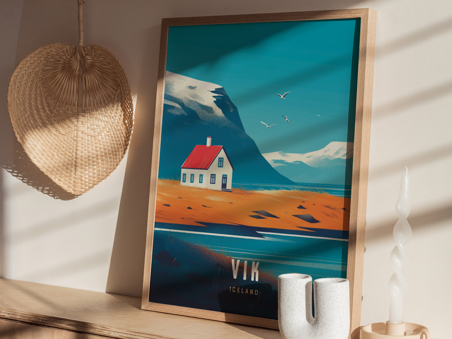 A framed poster of Vik, Iceland, featuring a coastal scene with a small house and birds flying over mountains. Its placed on a shelf with a woven basket and a.