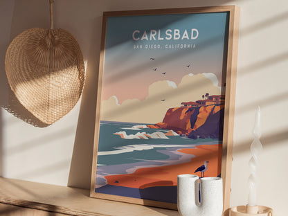 Framed poster of Carlsbad, California, depicting vibrant cliffs, ocean waves, and birds in the sky. It rests on a wooden shelf beside a woven wall decoration and modern candle.