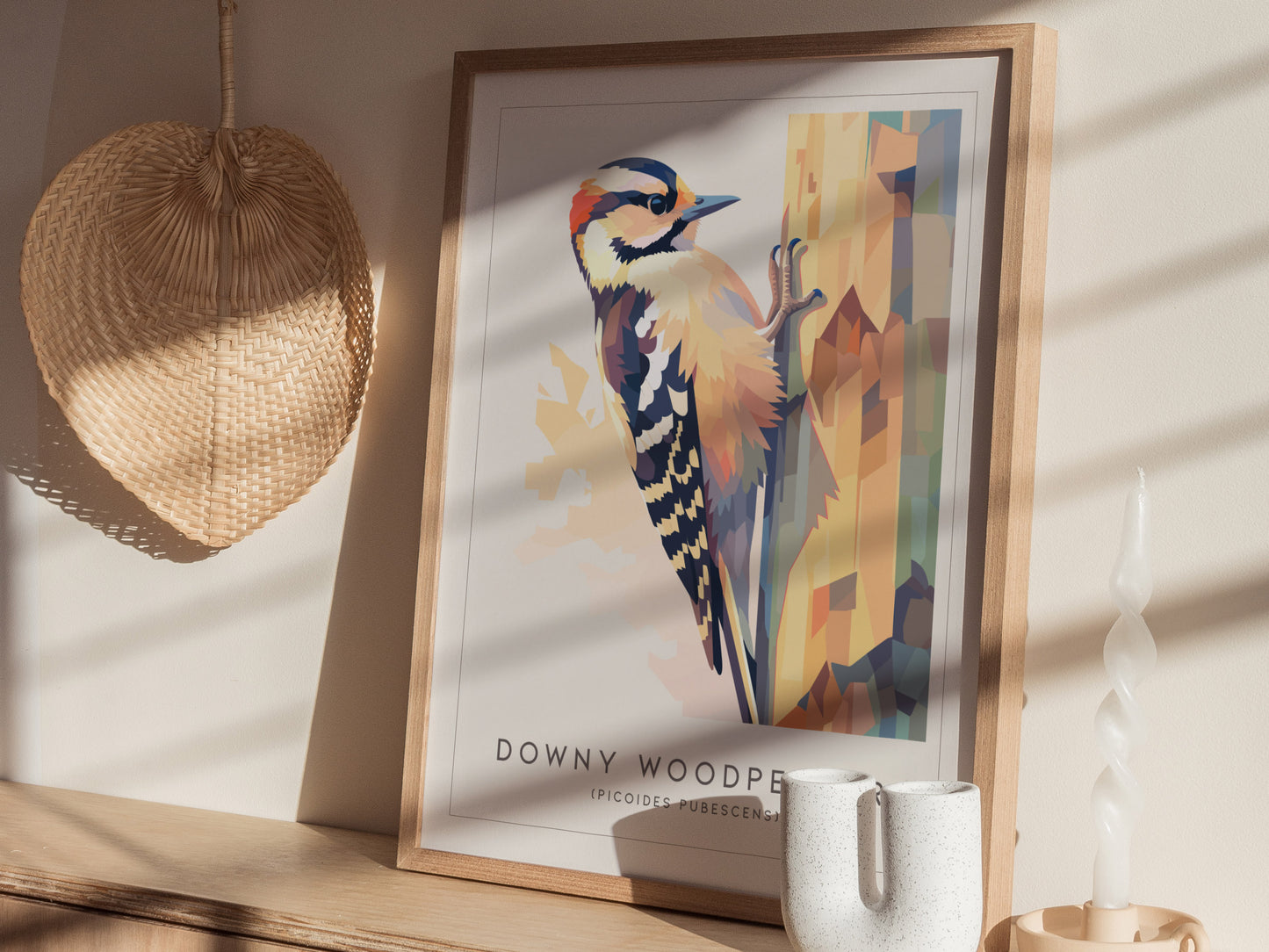 A framed art print of a Downy Woodpecker perched on a tree, showcasing vibrant geometric shapes and colors. The scene is illuminated by soft sunlight, with a woven basket and a decorative candle holder nearby, enhancing the cozy atmosphere.