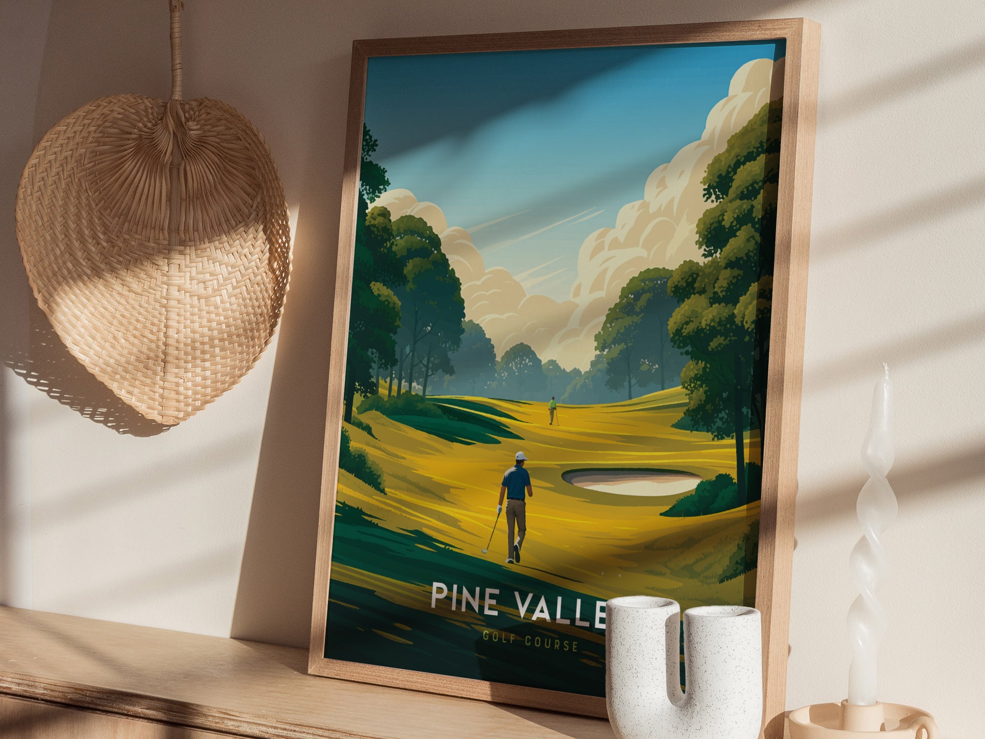 A framed Pine Valley Golf Course poster leans against a wall, depicting lush fairways and trees under a blue sky. A woven basket and abstract candle are nearby.