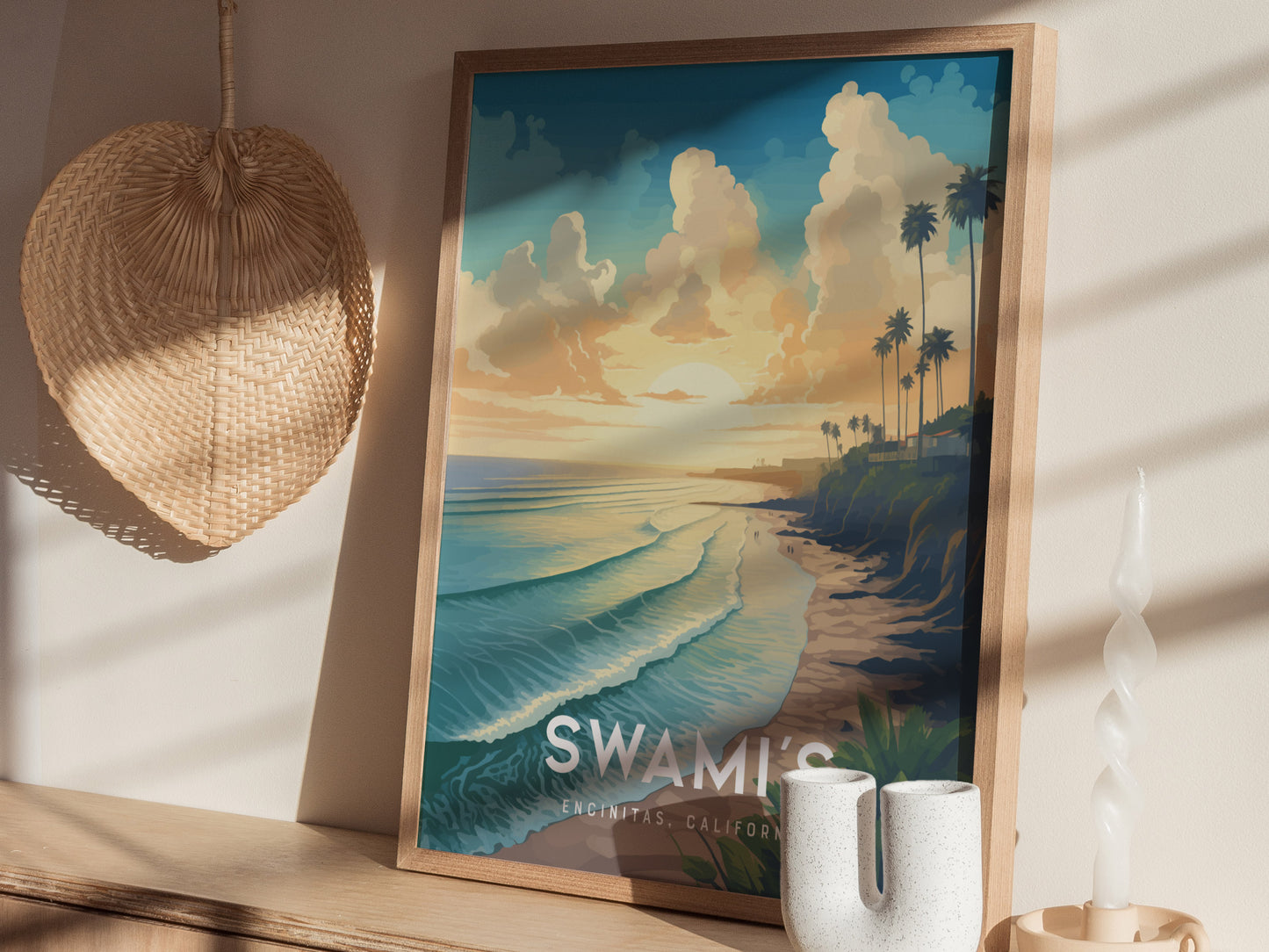 A framed poster of Swamis Encinitas Beach featuring a colorful sunset, palm trees, and waves is displayed on a shelf next to a woven hat and decorative candle.