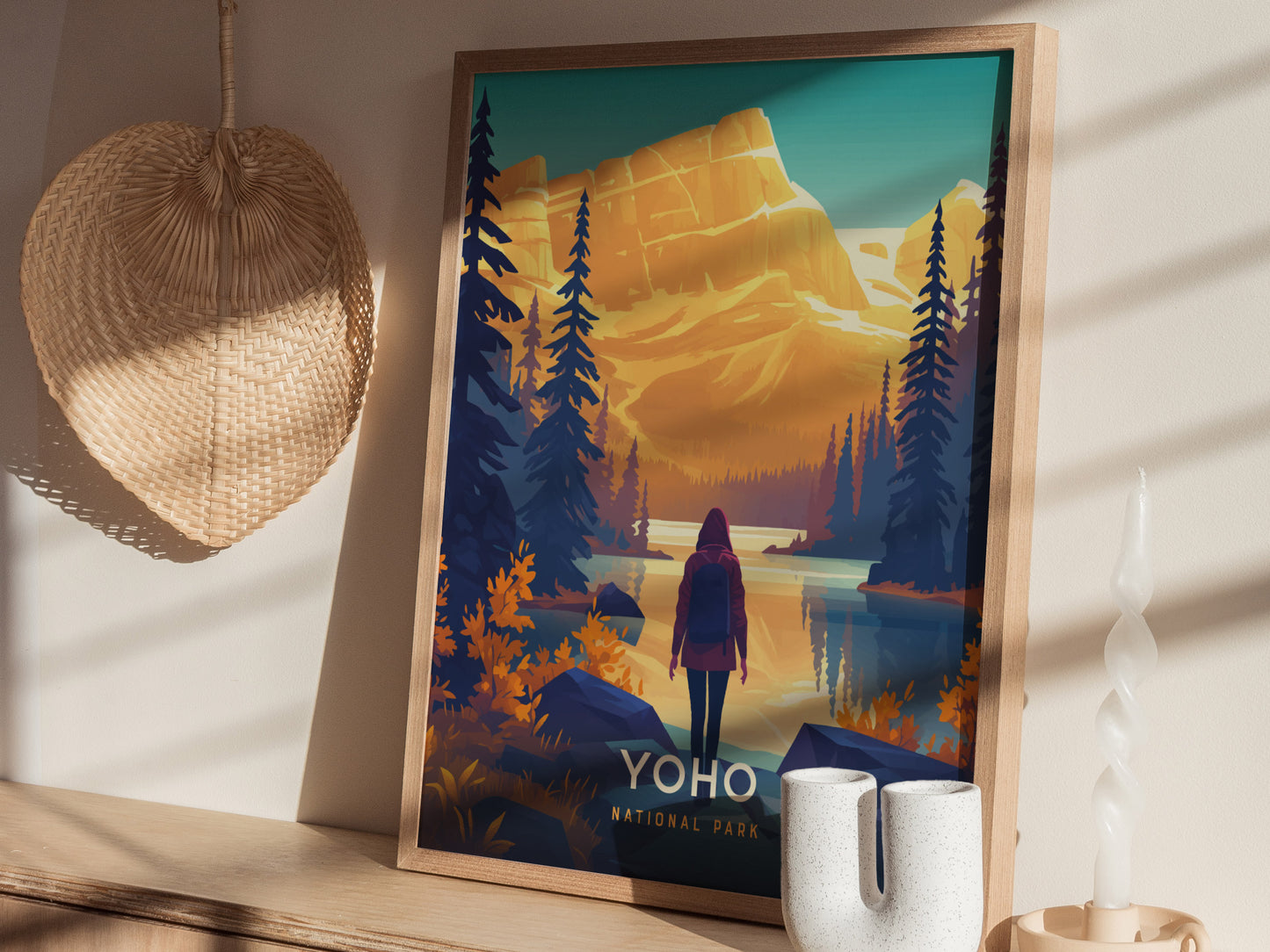 A framed poster of Yoho National Park features a person standing by a lake with pine trees and mountains. It is displayed on a wooden surface with a woven basket.