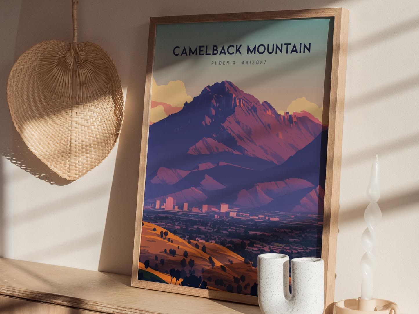 A framed Camelback Mountain poster rests on a wooden shelf, surrounded by a woven basket and decorative candle. Sunlight casts shadows across the poster and surrounding items.