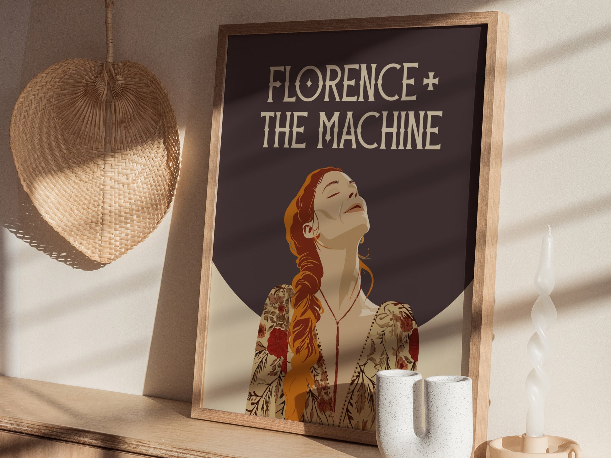 A framed Florence + The Machine poster featuring an illustrated figure with red hair is placed on a wooden surface, accompanied by a wicker fan and decorative candle.