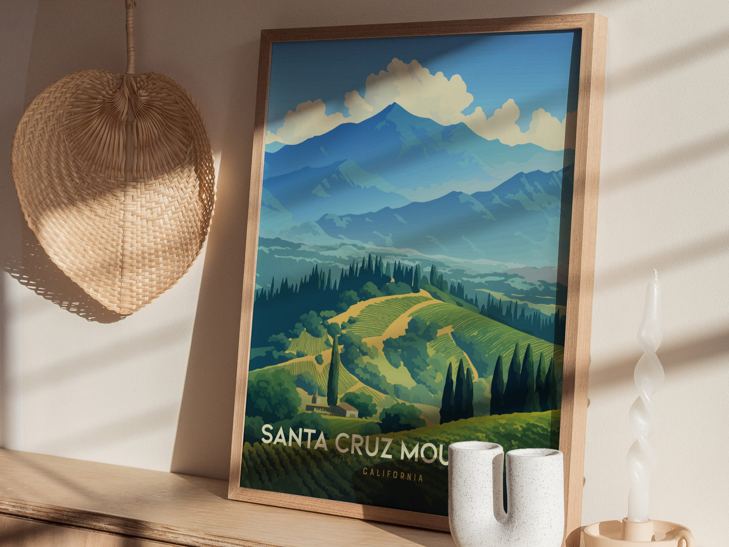 A framed poster of the Santa Cruz Mountains with lush vineyards and coastal peaks is displayed on a shelf beside a woven hat and a white ceramic vase.
