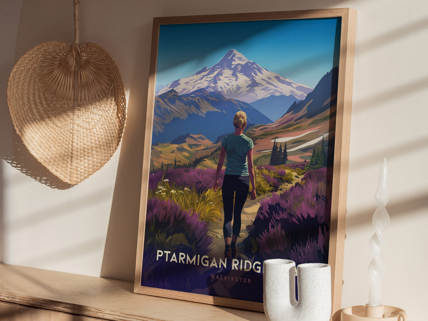 A framed poster depicting a hiker on Ptarmigan Ridge with Mt. Baker in the background is displayed on a wooden shelf, alongside a woven basket and white ceramic candle holder.