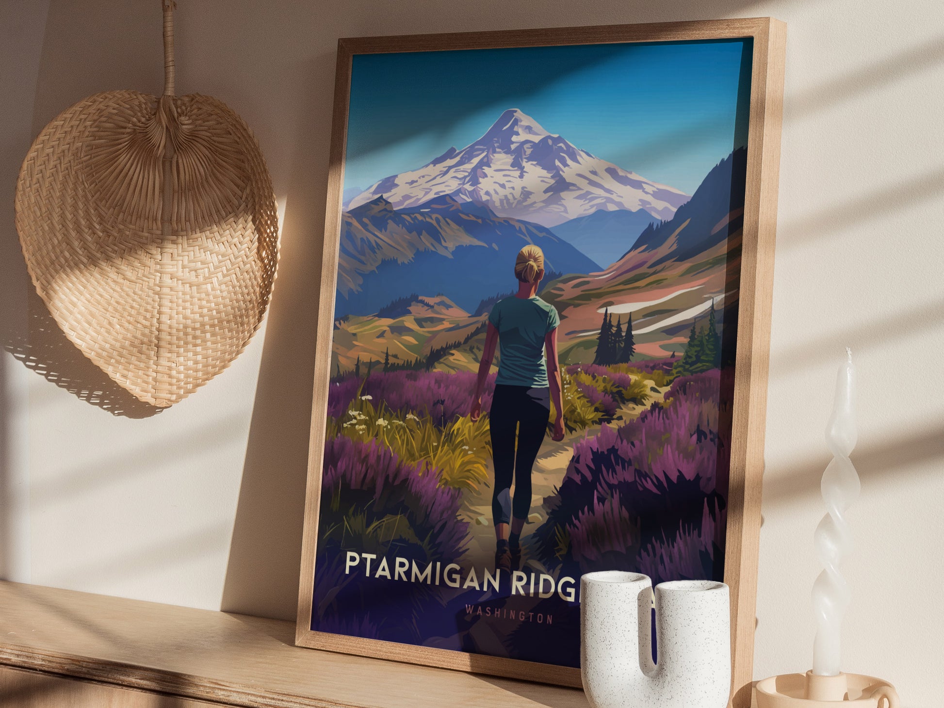 A framed poster depicting a hiker on Ptarmigan Ridge with Mt. Baker in the background is displayed on a wooden shelf, alongside a woven basket and white ceramic candle holder.