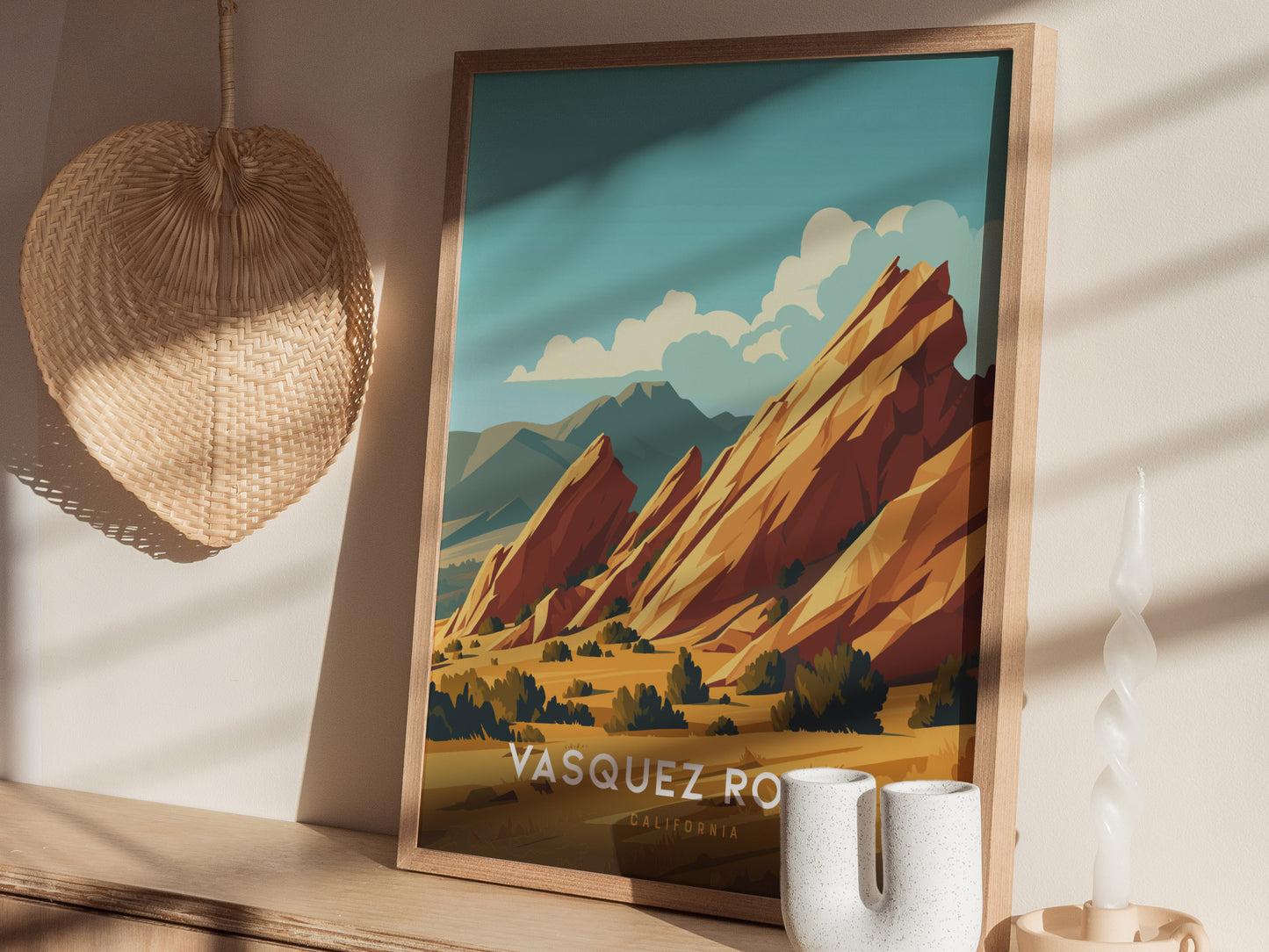 A framed poster of Vasquez Rocks with tilted sandstone formations sits on a wooden shelf next to a woven wall decoration and a spiral candle.