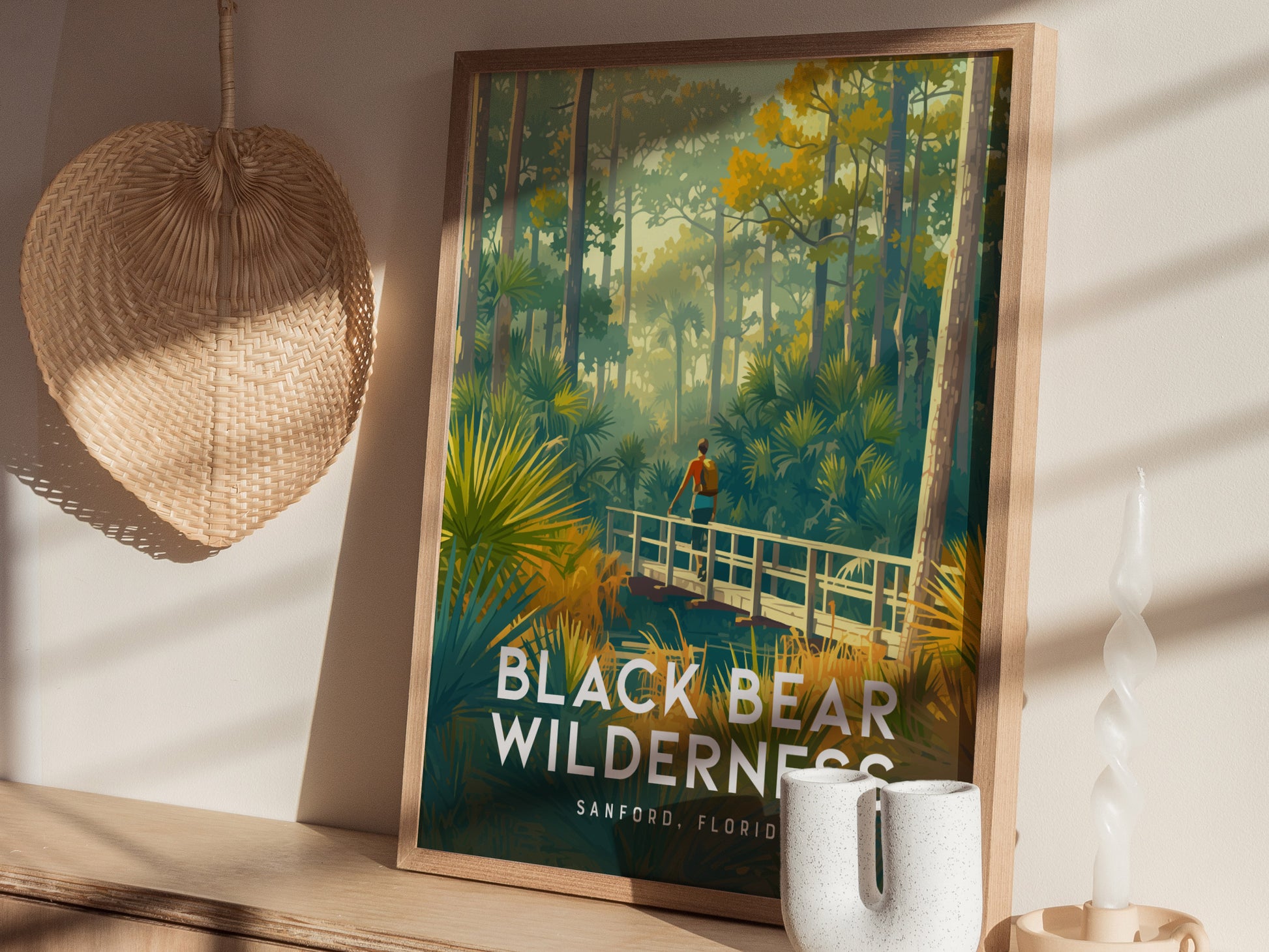 A framed poster of a hiker on a wooden bridge in a lush forest, labeled Black Bear Wilderness, Sanford, Florida, is displayed with a woven fan and decorative items.