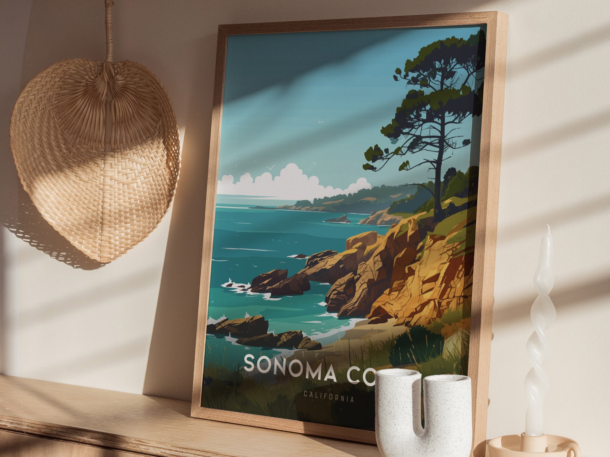 A framed poster of the Sonoma Coast with sea cliffs and a tree is displayed on a wooden shelf alongside a woven decor piece and a twisted white candle.