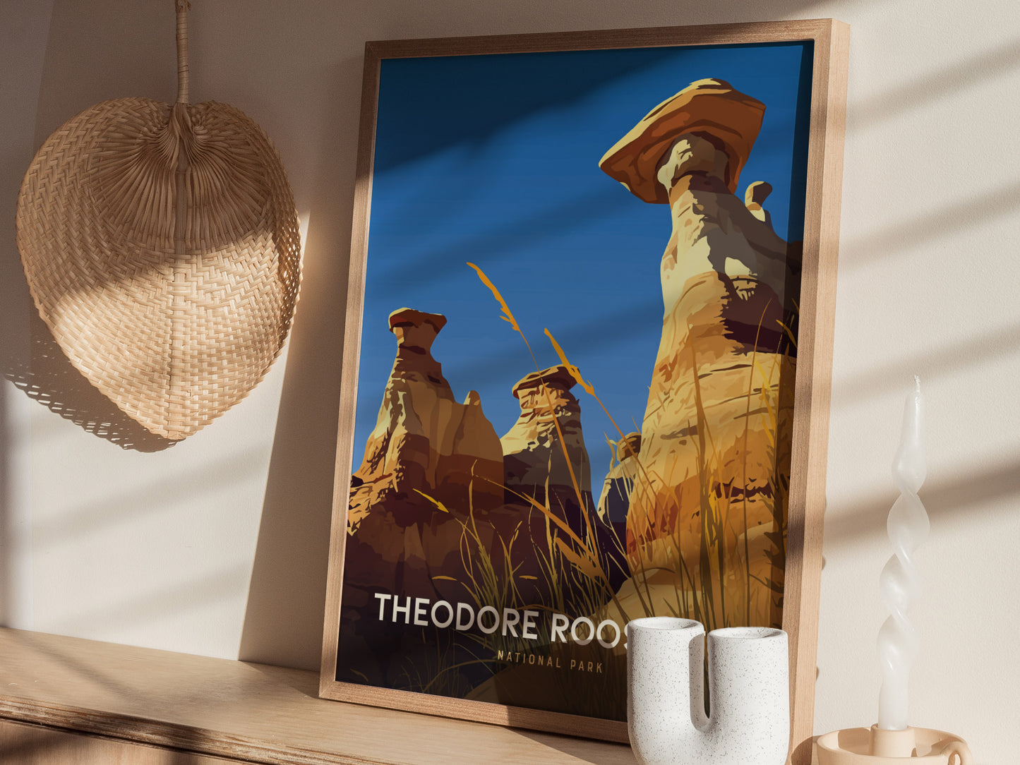 A framed poster of Theodore Roosevelt National Park features dramatic hoodoos and a blue sky. Its displayed on a wooden shelf alongside a woven fan and a twisted candle.
