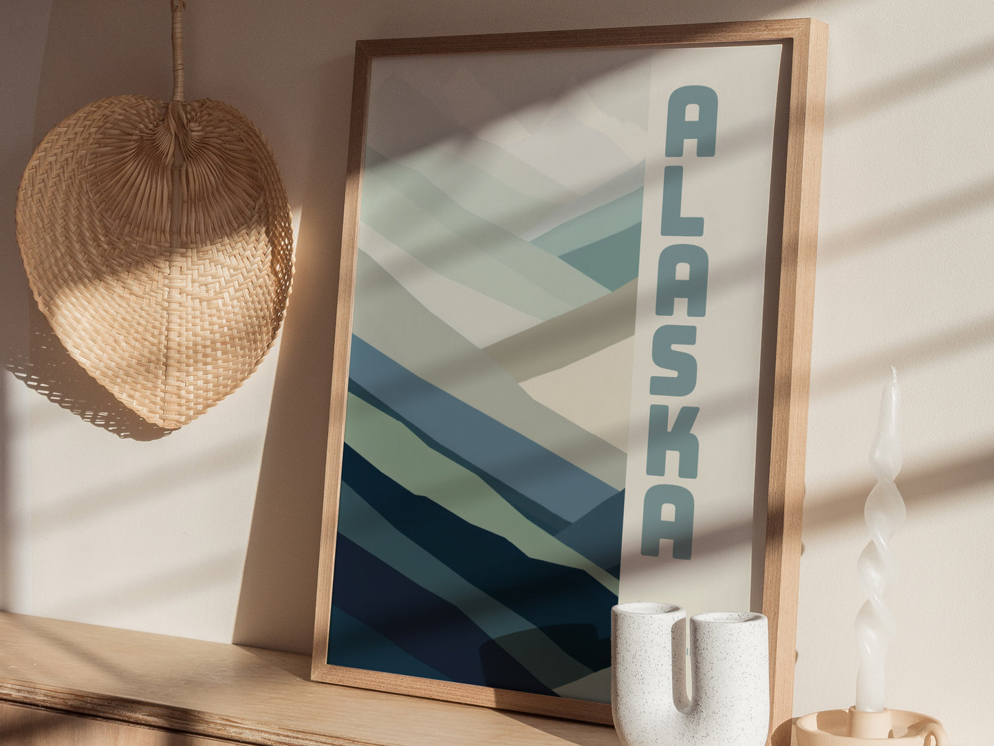 A framed minimalist poster with ALASKA text and abstract blue and green shapes, placed on a wooden surface with a woven basket and a twisted white candle nearby.