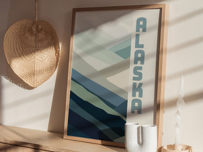 A framed minimalist poster with ALASKA text and abstract blue and green shapes, placed on a wooden surface with a woven basket and a twisted white candle nearby.