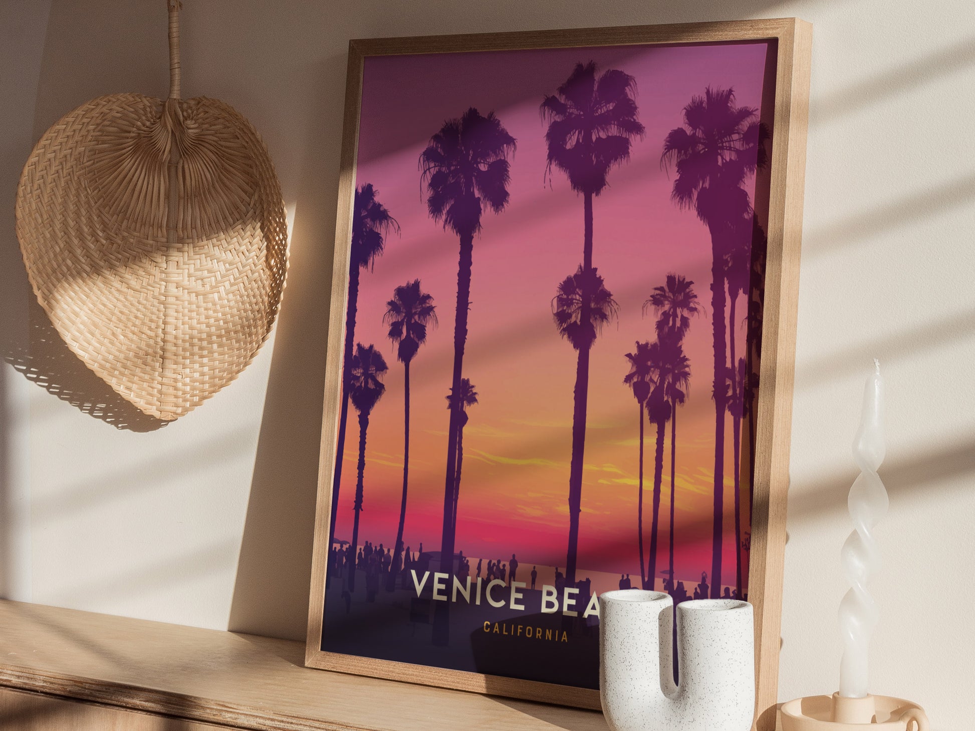 A framed poster depicting a vibrant Venice Beach sunset with silhouetted palm trees, set on a wooden shelf next to a woven basket, candle, and ceramic vase.