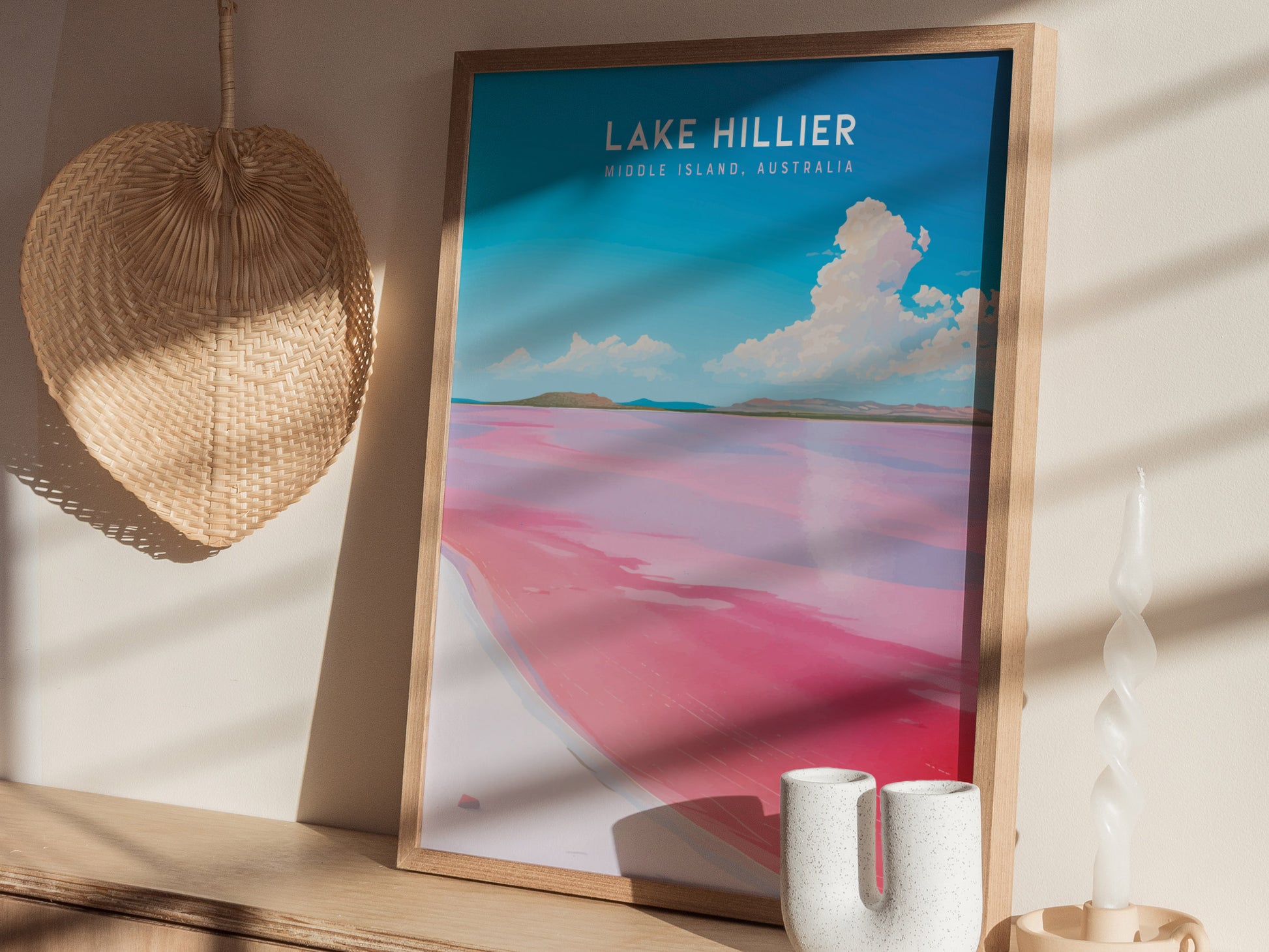 A framed poster of Lake Hillier, showcasing vibrant pink water against a blue sky, is displayed on a wooden shelf with a woven fan and decorative candle nearby.