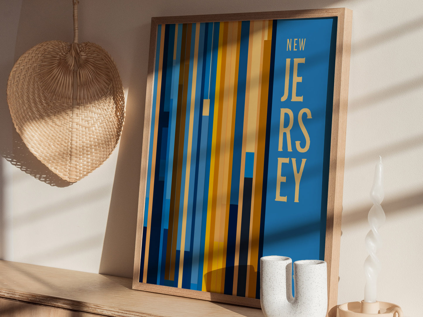 A colorful framed poster with bold stripes and the text New Jersey leans against a wall. Nearby are a woven straw fan and a twisted white candle.