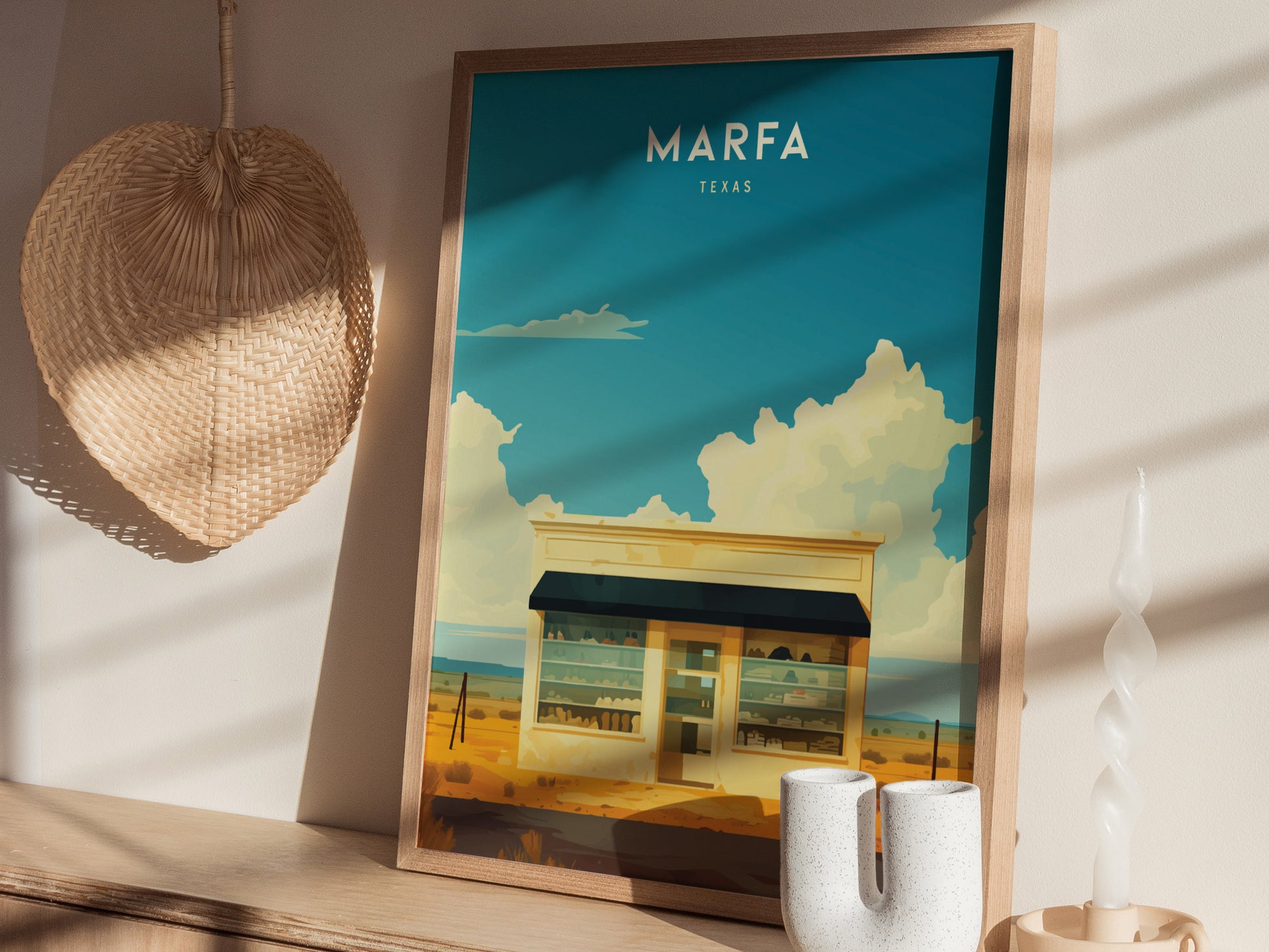 A framed Marfa Texas poster depicting a desert scene with clouds is displayed on a wooden surface, next to a wicker wall decoration and a twisted candle.