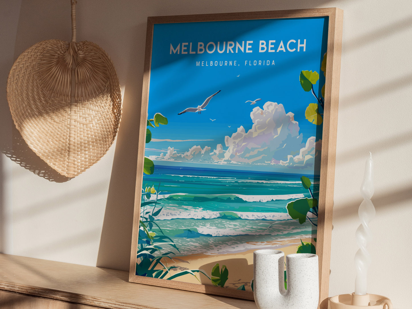 A framed poster of Melbourne Beach in vibrant colors shows seagulls, ocean waves, and clouds. Its displayed on a wooden shelf with a woven hat, candle, and ceramic vase.