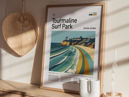Framed Tourmaline Surf Park poster with vibrant beach artwork rests against a wall, alongside a woven fan and a twisted candle. Sunlight casts soft shadows on the display.