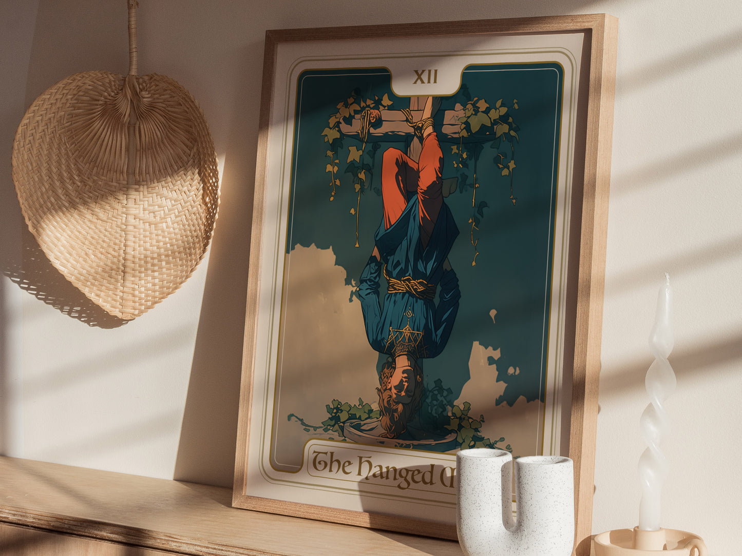 A framed Hanged Man tarot card poster is displayed on a wooden shelf, with a wicker fan and a twisted white candle nearby, casting soft shadows.
