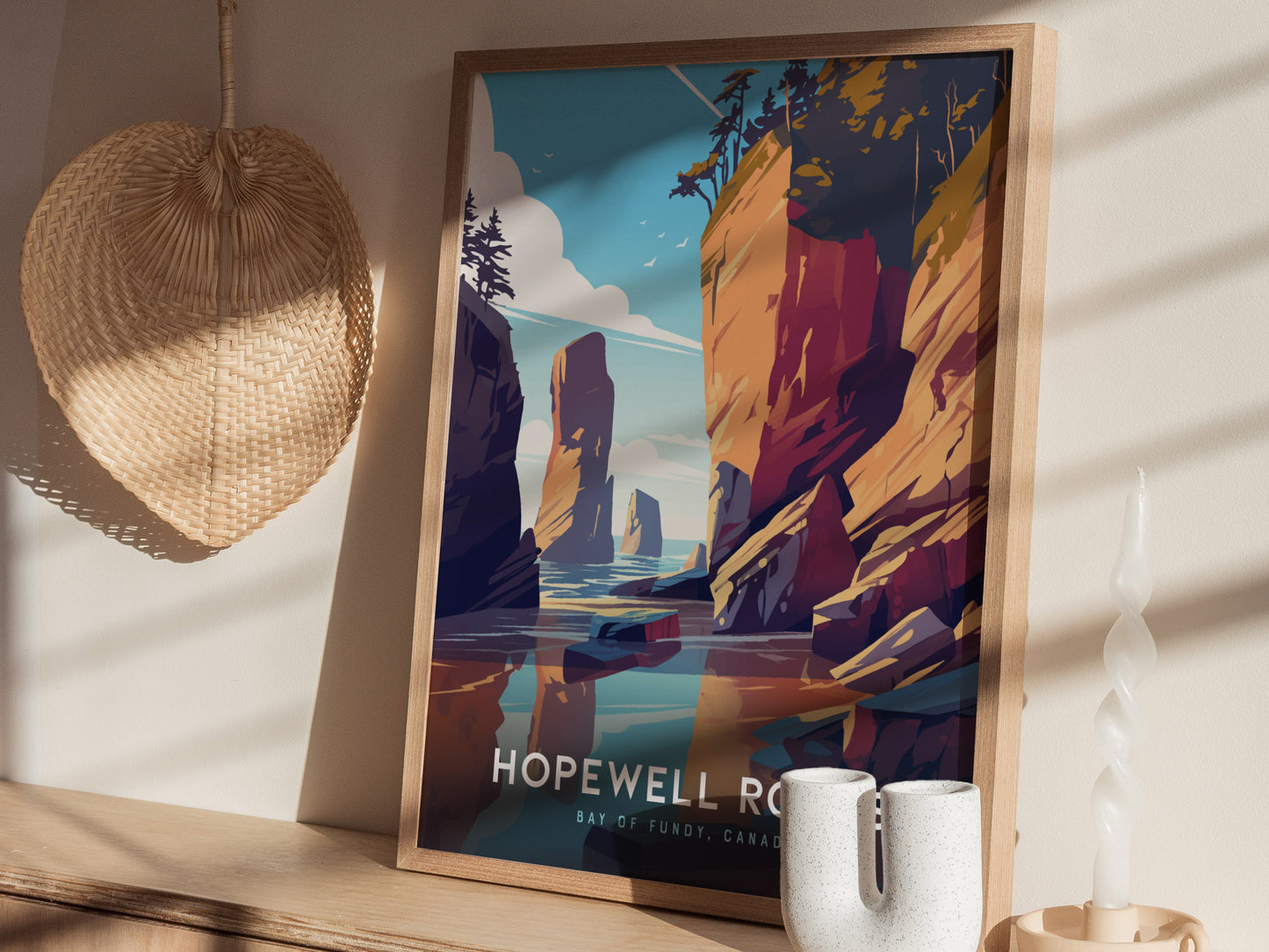A framed poster of Hopewell Rocks depicting vibrant sandstone cliffs and blue waters is displayed on a wooden shelf with a woven basket and decorative candle nearby.