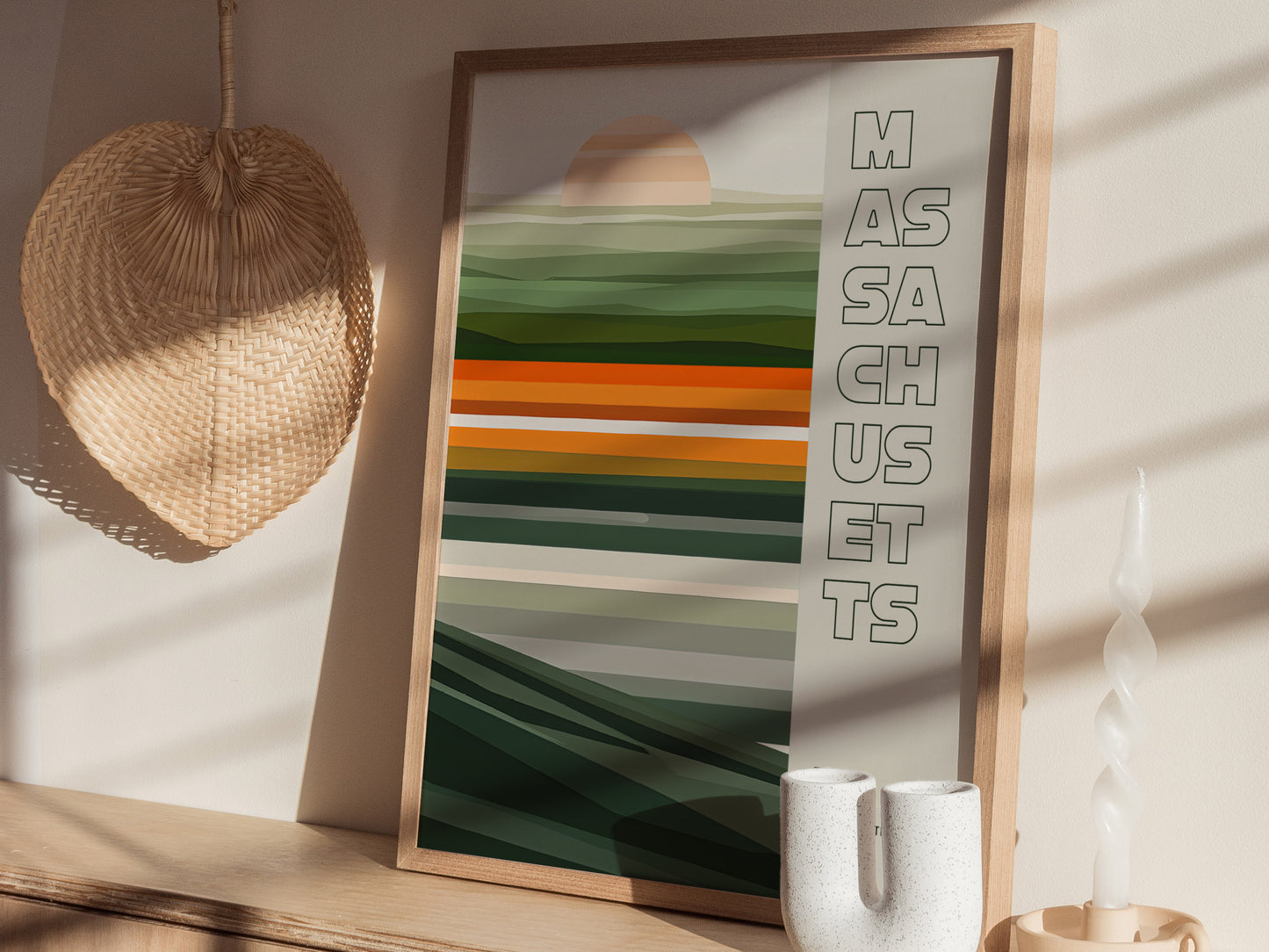 A framed poster with abstract green and orange stripes and the word Massachusetts is displayed on a wooden shelf, alongside a woven wall decoration and a white twisted candle.