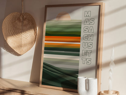A framed poster with abstract green and orange stripes and the word Massachusetts is displayed on a wooden shelf, alongside a woven wall decoration and a white twisted candle.