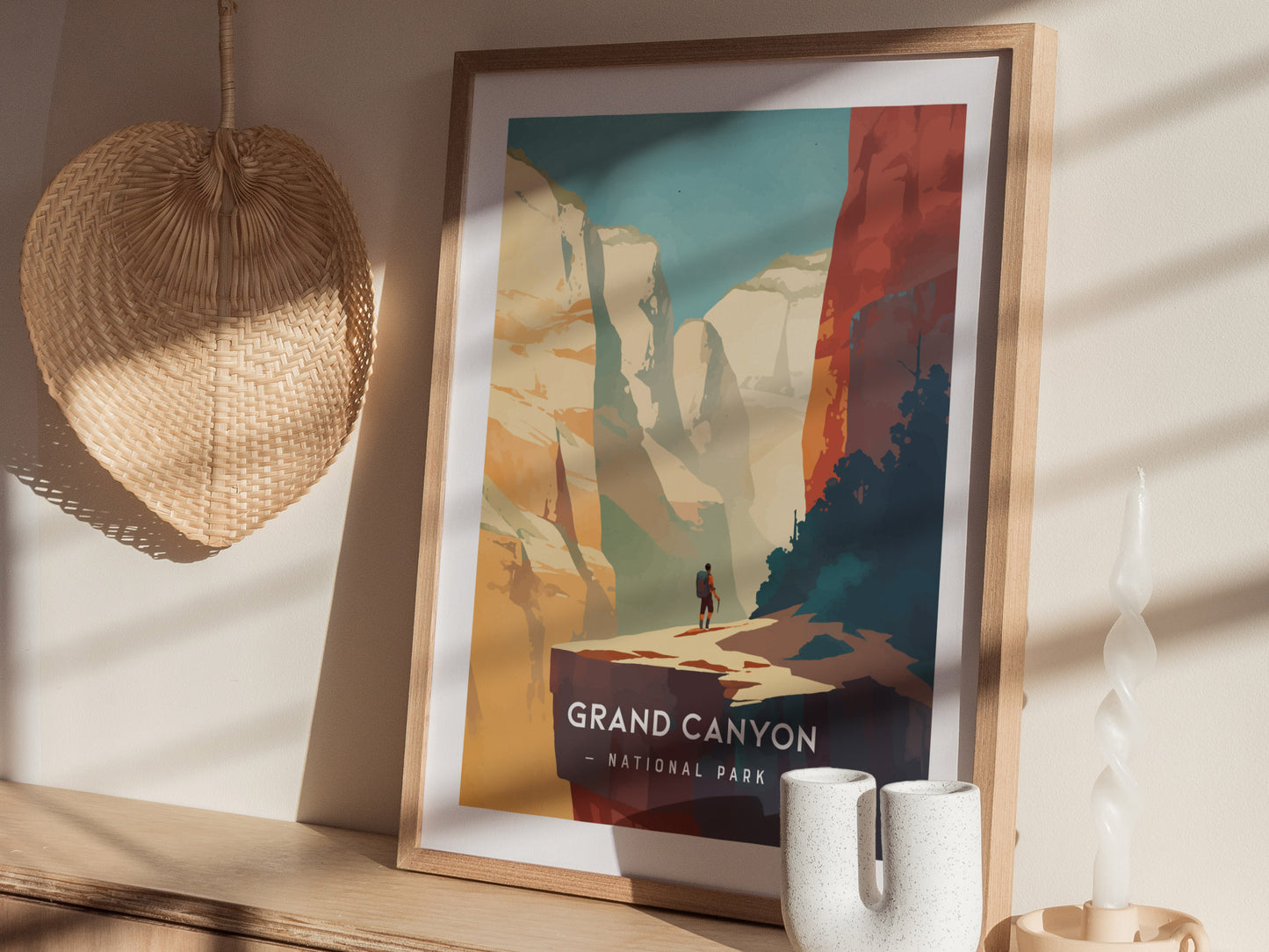 A framed Grand Canyon National Park poster with a minimalist design is displayed on a wooden surface, alongside a woven basket and a twisted candle.