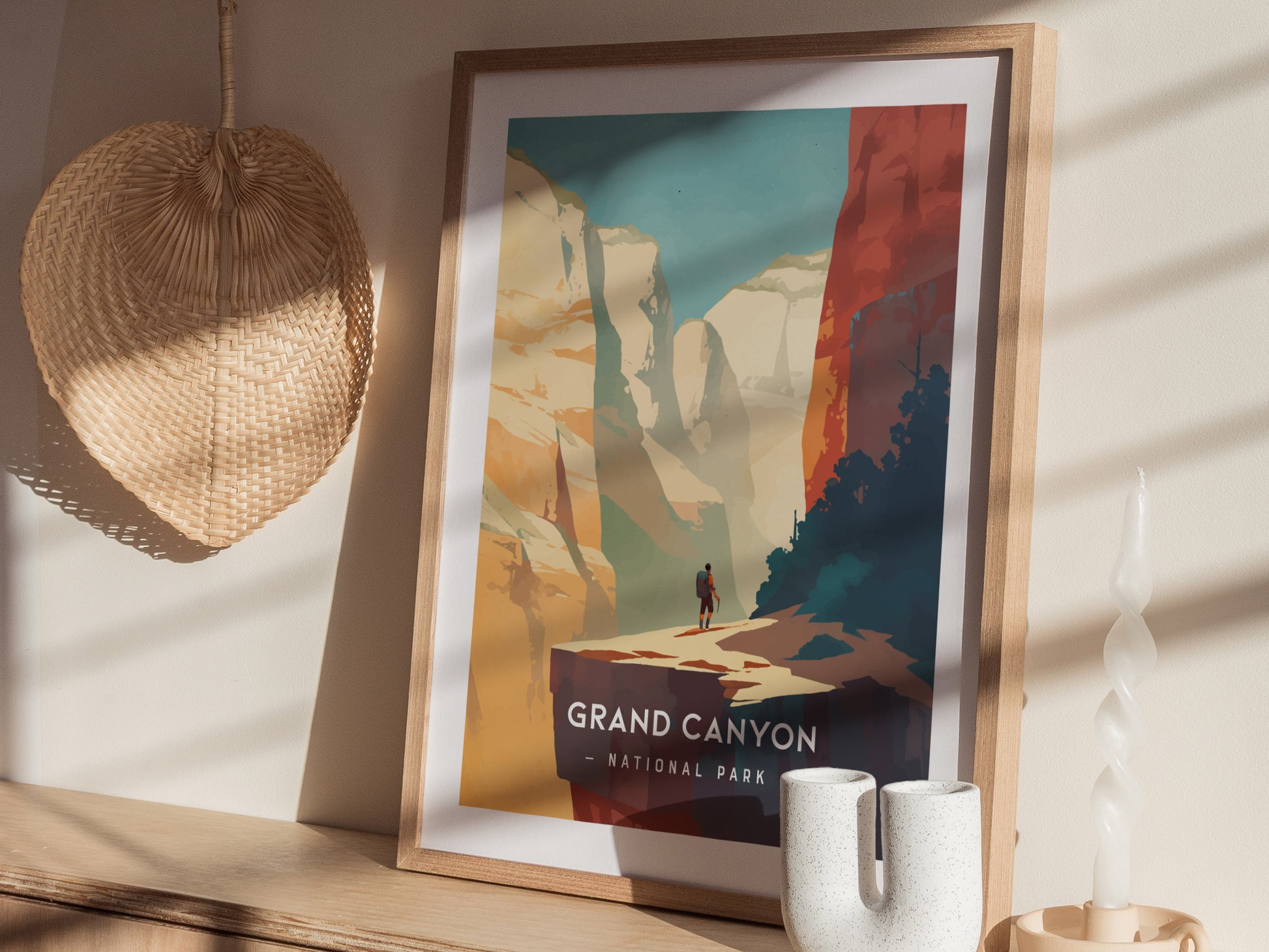 A framed Grand Canyon National Park poster with a minimalist design is displayed on a wooden surface, alongside a woven basket and a twisted candle.