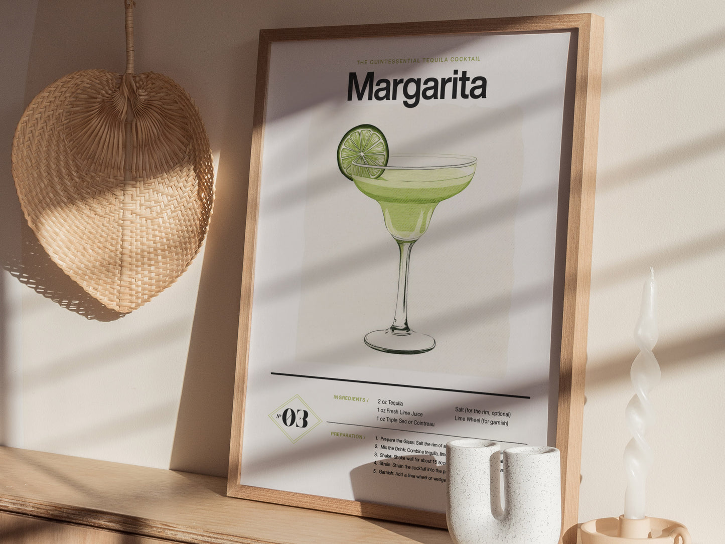 A framed Margarita cocktail poster with an illustrated drink and lime slice sits on a wooden shelf. Nearby, a woven fan and twisted candle add decorative elements.