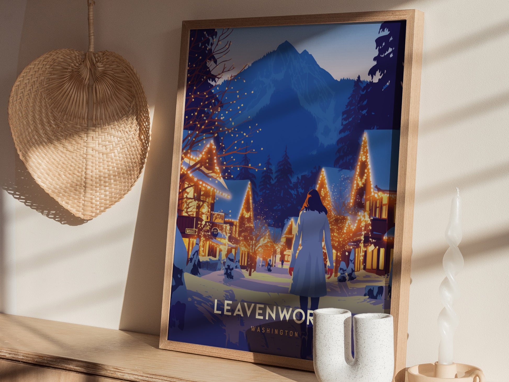 A framed poster of Leavenworth, Washington, showing a winter scene with a person in a coat walking through a village decorated with warm lights and snowy mountains in the background.