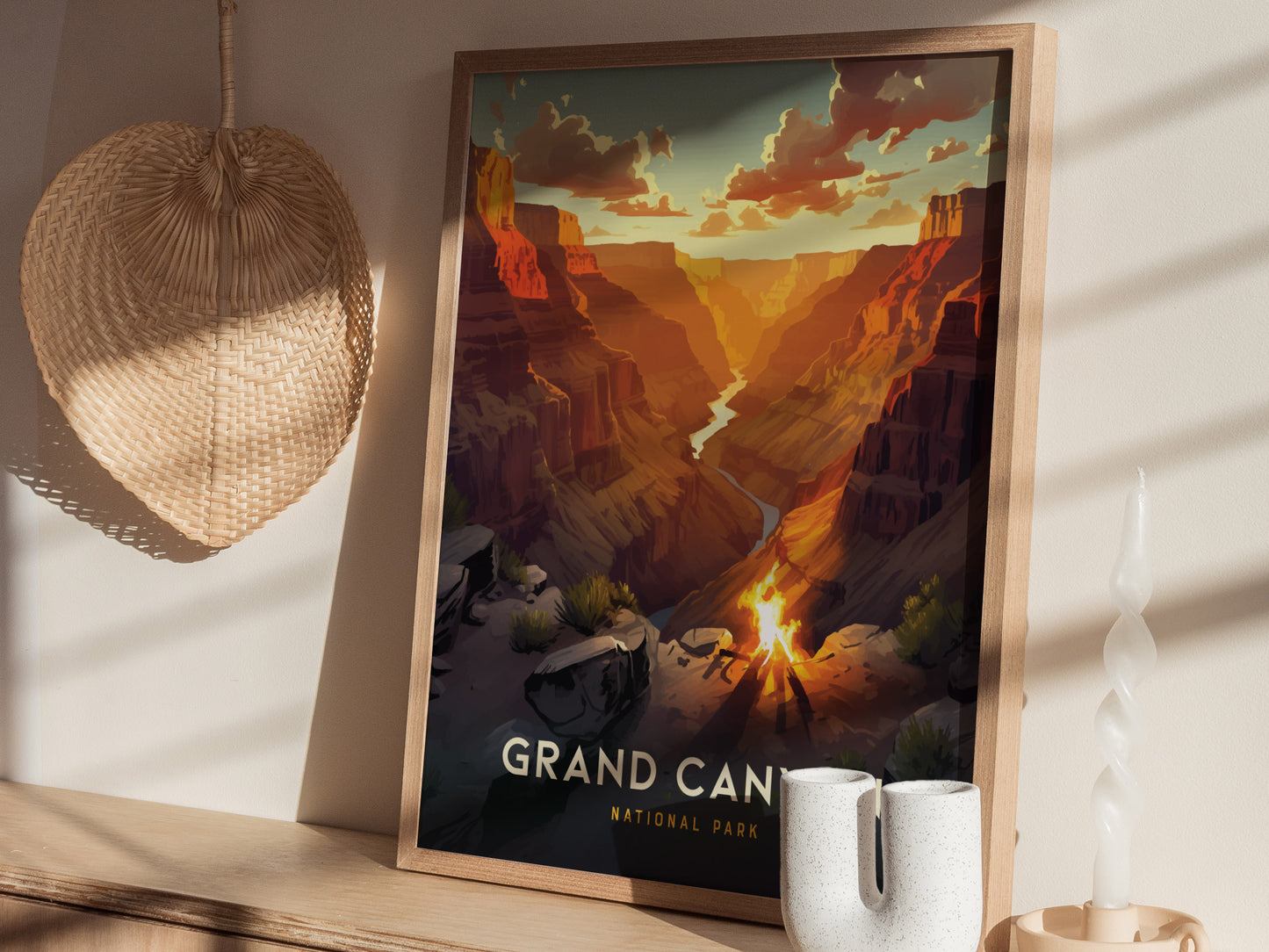 Framed poster of the Grand Canyon with vibrant orange and red hues, displayed on a wooden surface alongside a wicker fan, ceramic vase, and twisted candle.