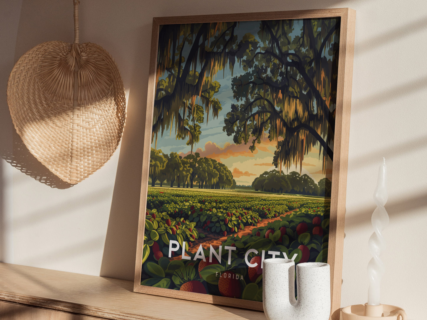 A framed poster of Plant City, Florida, features vibrant strawberry fields under a sunrise with oak trees and Spanish moss. Its displayed on a shelf alongside a wicker fan and.