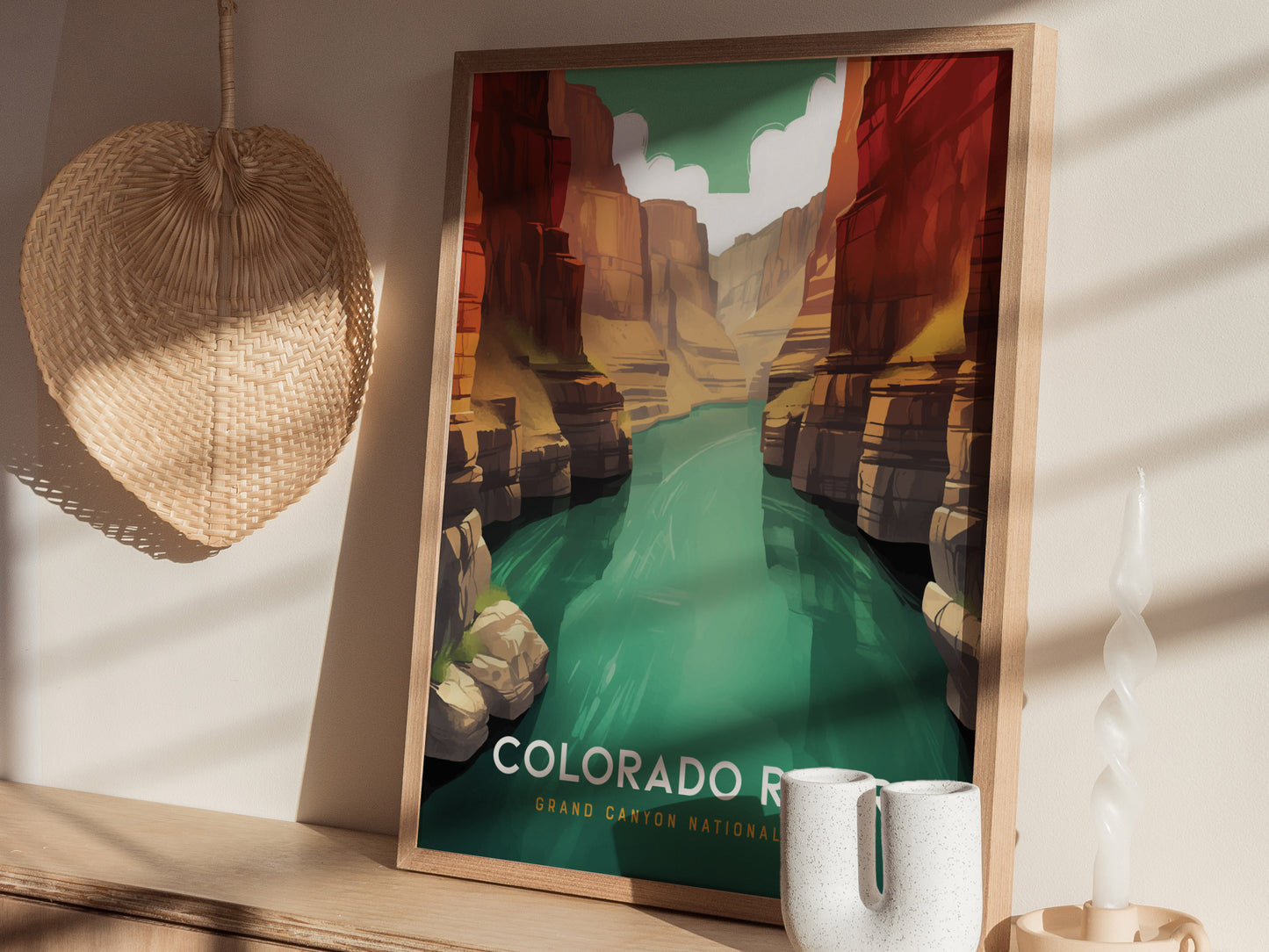 A framed poster of the Colorado River in the Grand Canyon is displayed on a wooden shelf, accompanied by a woven basket and a twisted white candle.
