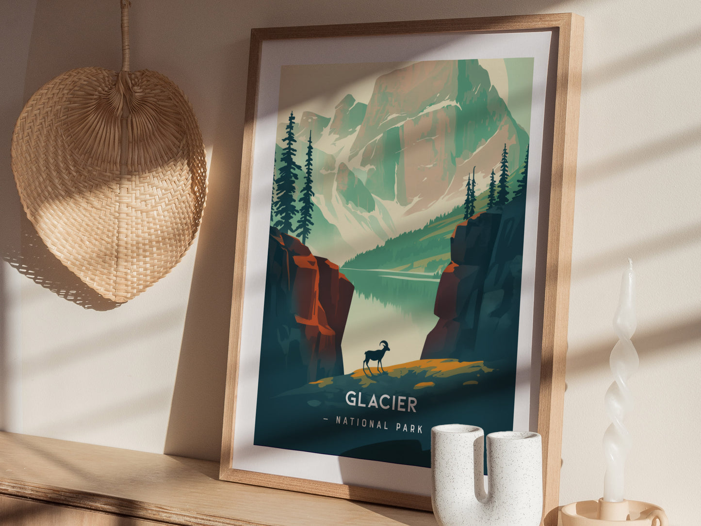 A framed minimalist poster of Glacier National Park features a serene alpine lake, rocky cliffs, and a lone bighorn sheep. Its displayed on a wooden shelf beside a woven fan.