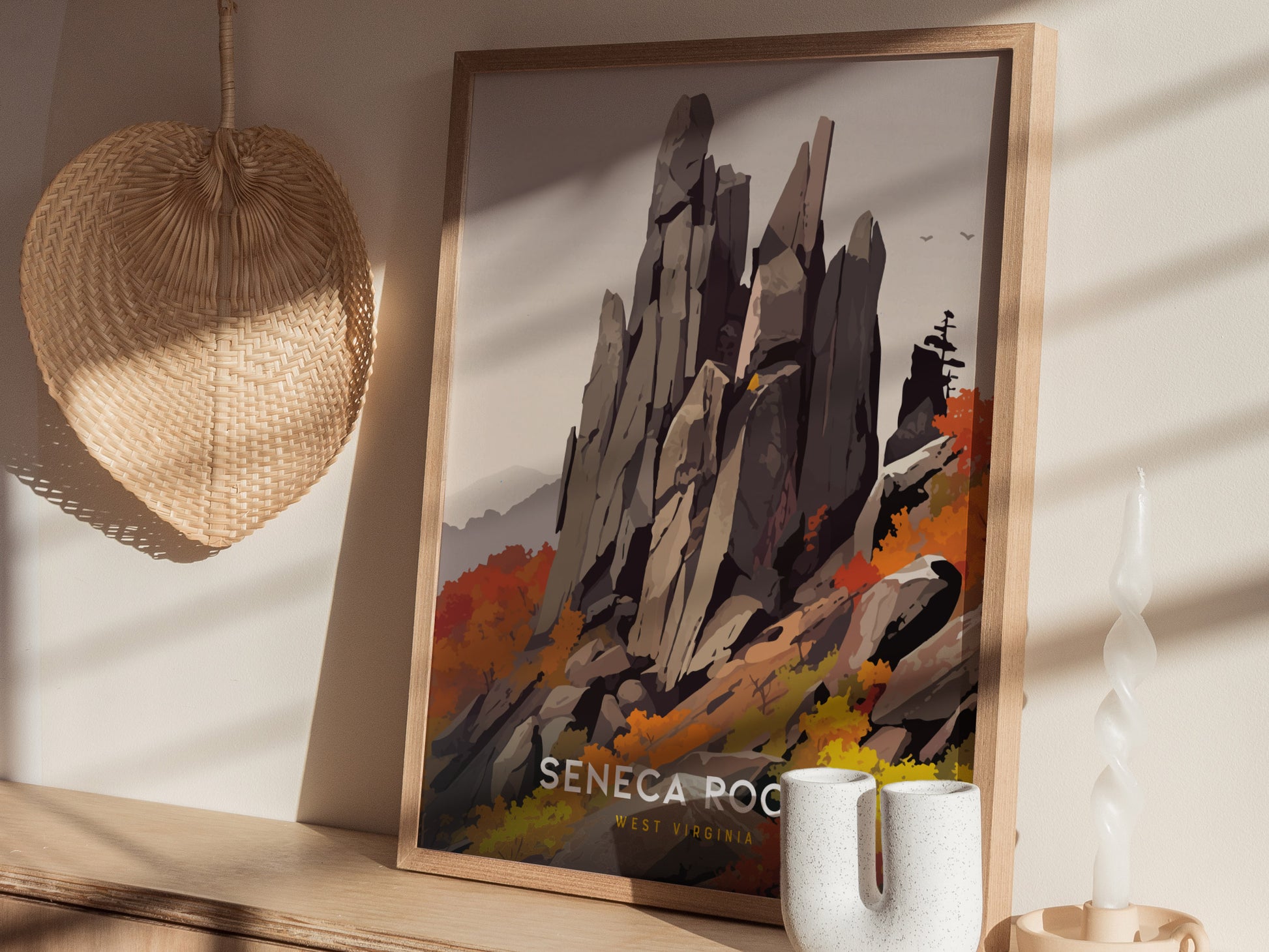 A framed poster of Seneca Rocks, featuring towering rock formations and vibrant autumn foliage, is displayed on a wooden surface beside a woven fan and a twisted candle.