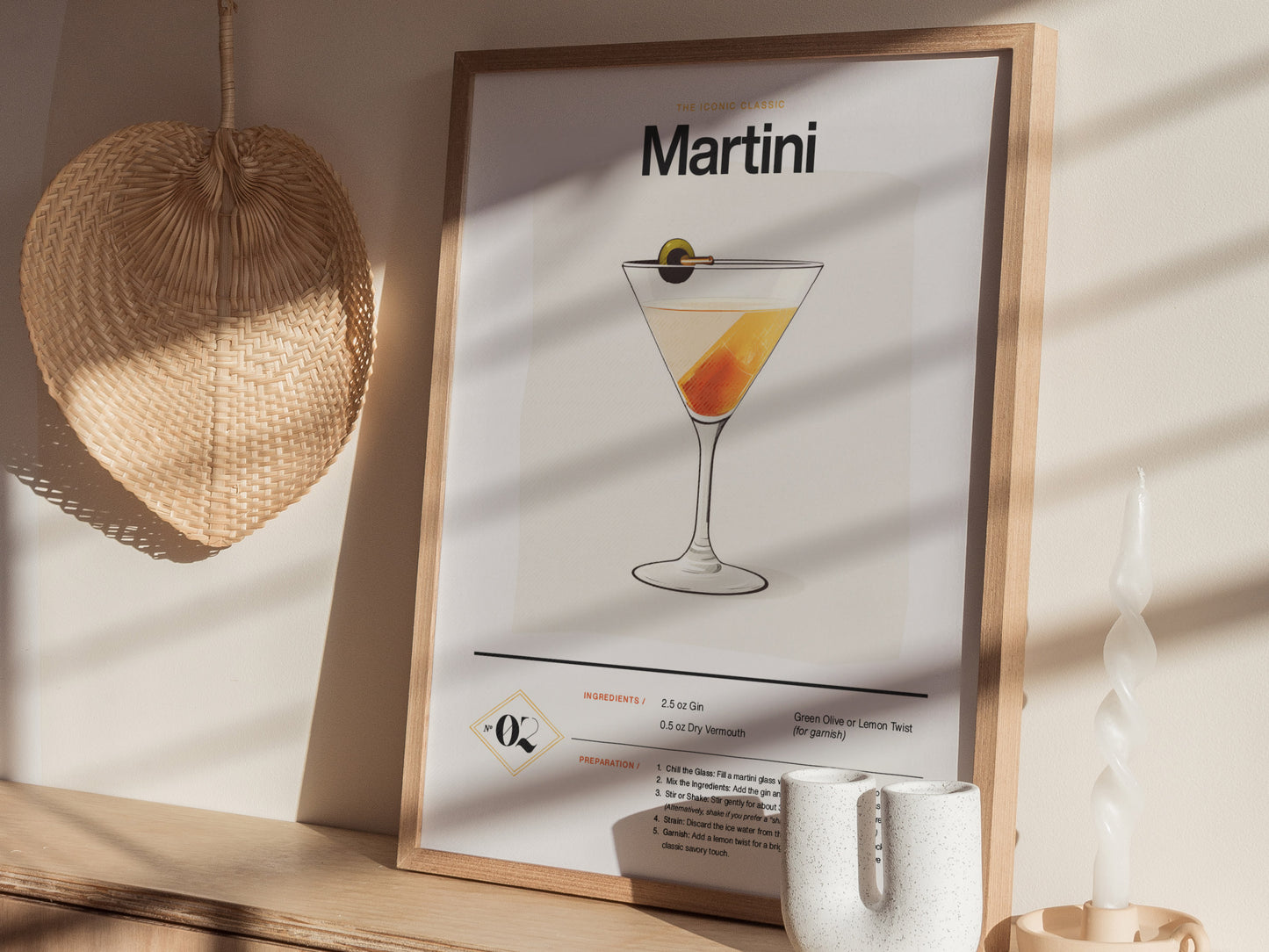 A framed poster of a martini cocktail recipe is displayed on a wooden shelf next to a woven fan and a twisted white candle. Sunlight casts shadows on the wall.