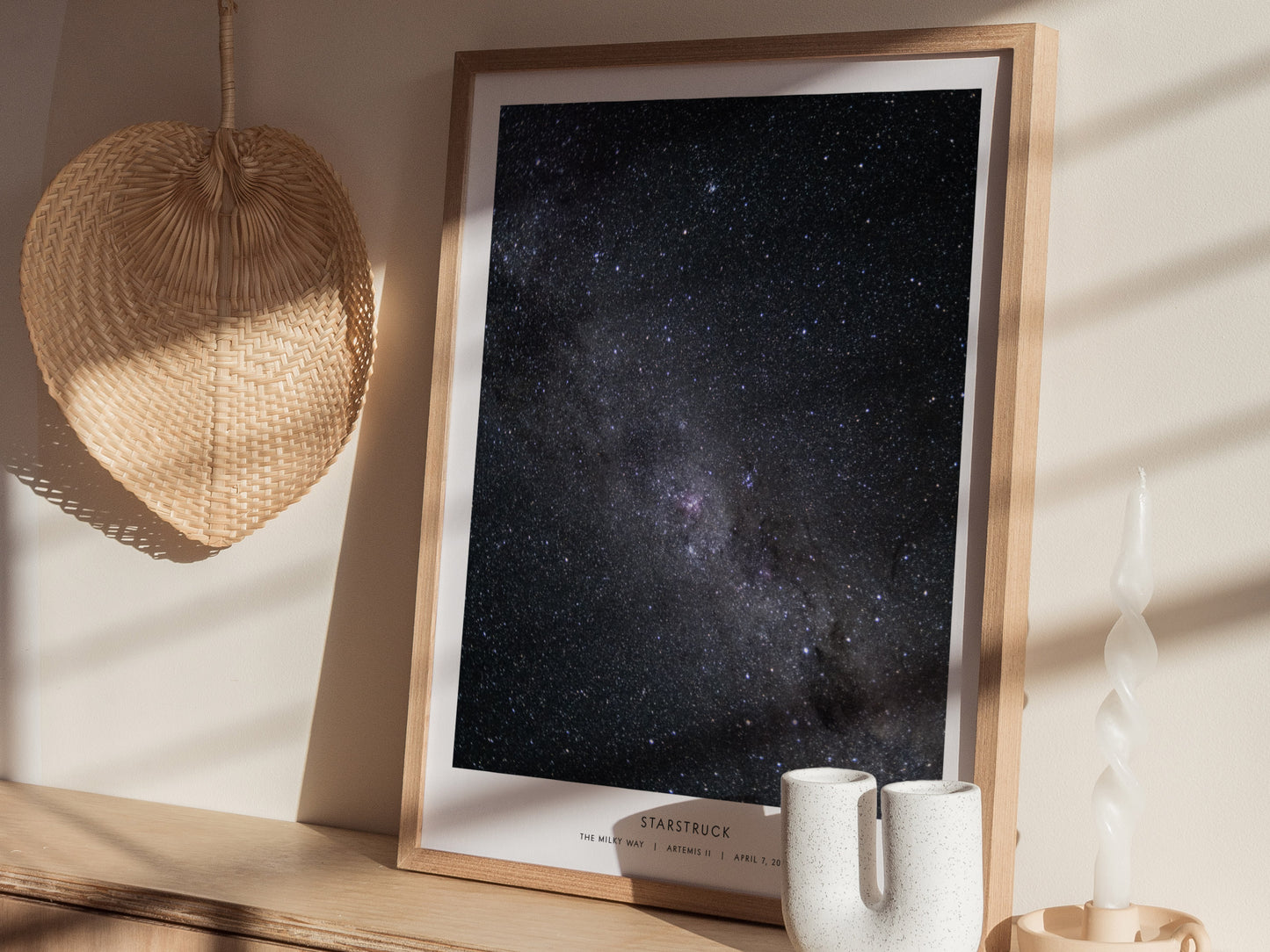 A framed art print titled Starstruck is displayed on a wooden table. The poster features a stunning view of a starry night sky, showcasing countless stars against a dark backdrop, with soft sunlight filtering through nearby objects,.