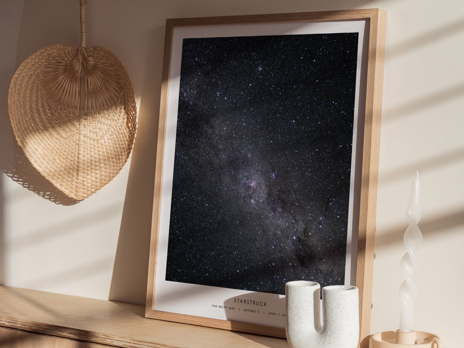 A framed art print titled Starstruck is displayed on a wooden table. The poster features a stunning view of a starry night sky, showcasing countless stars against a dark backdrop, with soft sunlight filtering through nearby objects,.