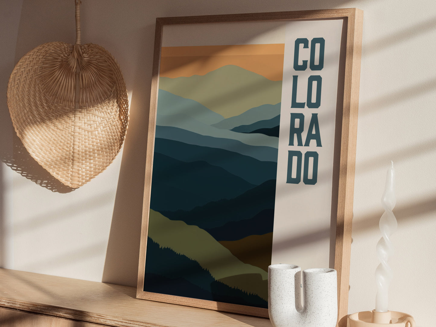 A framed minimalist poster depicting abstract Rocky Mountains with layered hues, alongside the word Colorado. It rests on a wooden shelf with a woven basket and a twisted candle.