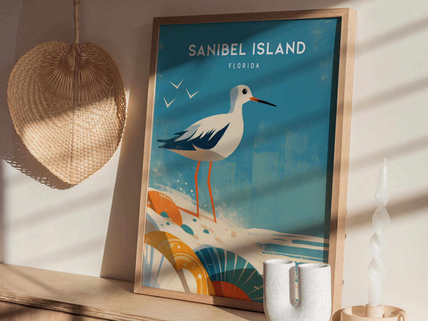 A framed Sanibel Island poster featuring a sandpiper and seashells is displayed on a wooden surface, next to a wicker fan and a twisted white candle.