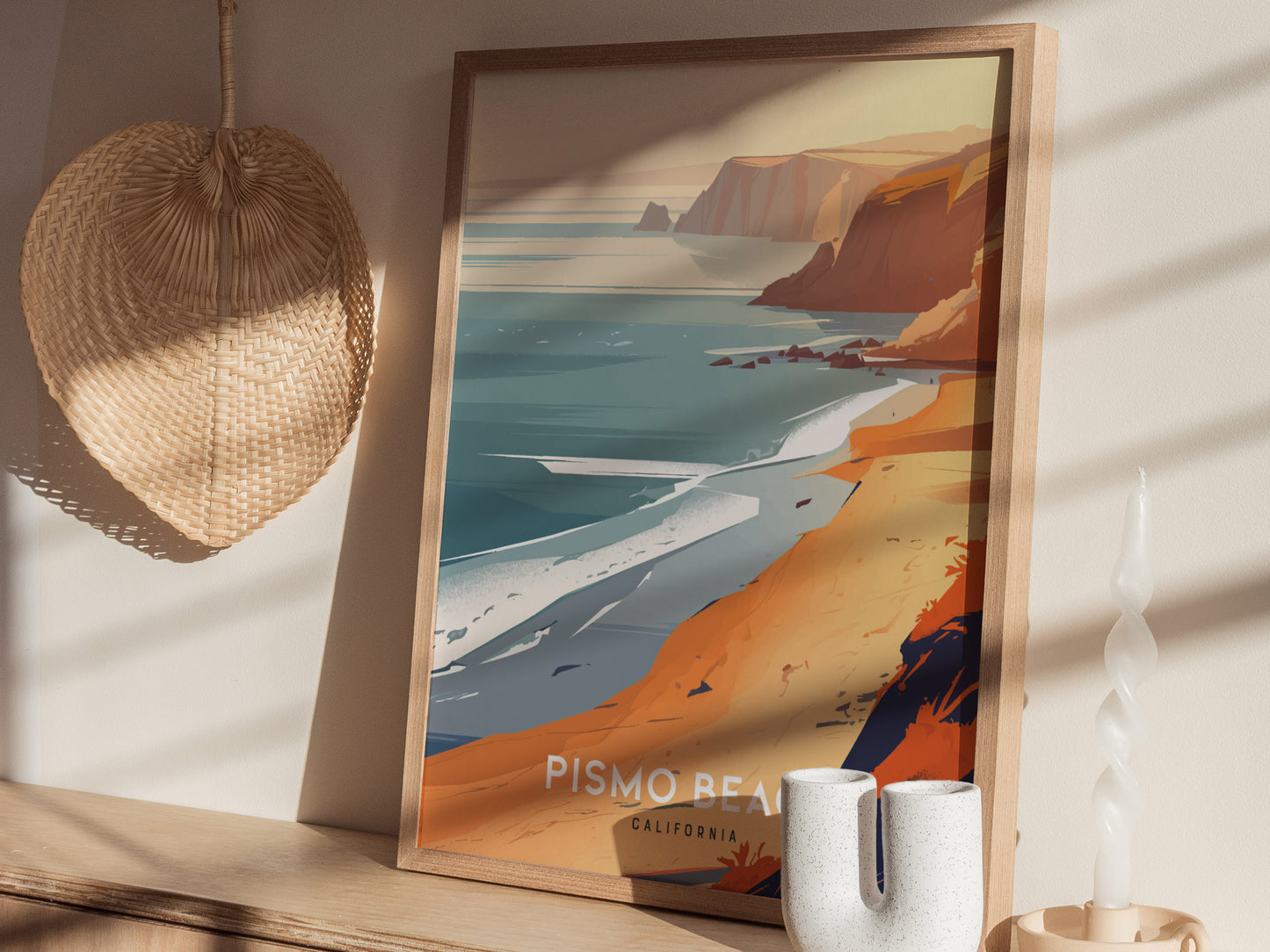 Framed artwork of Pismo Beach showcasing cliffs and ocean, placed on a wooden shelf with a woven fan and decorative candle nearby. Soft shadows enhance the serene atmosphere.