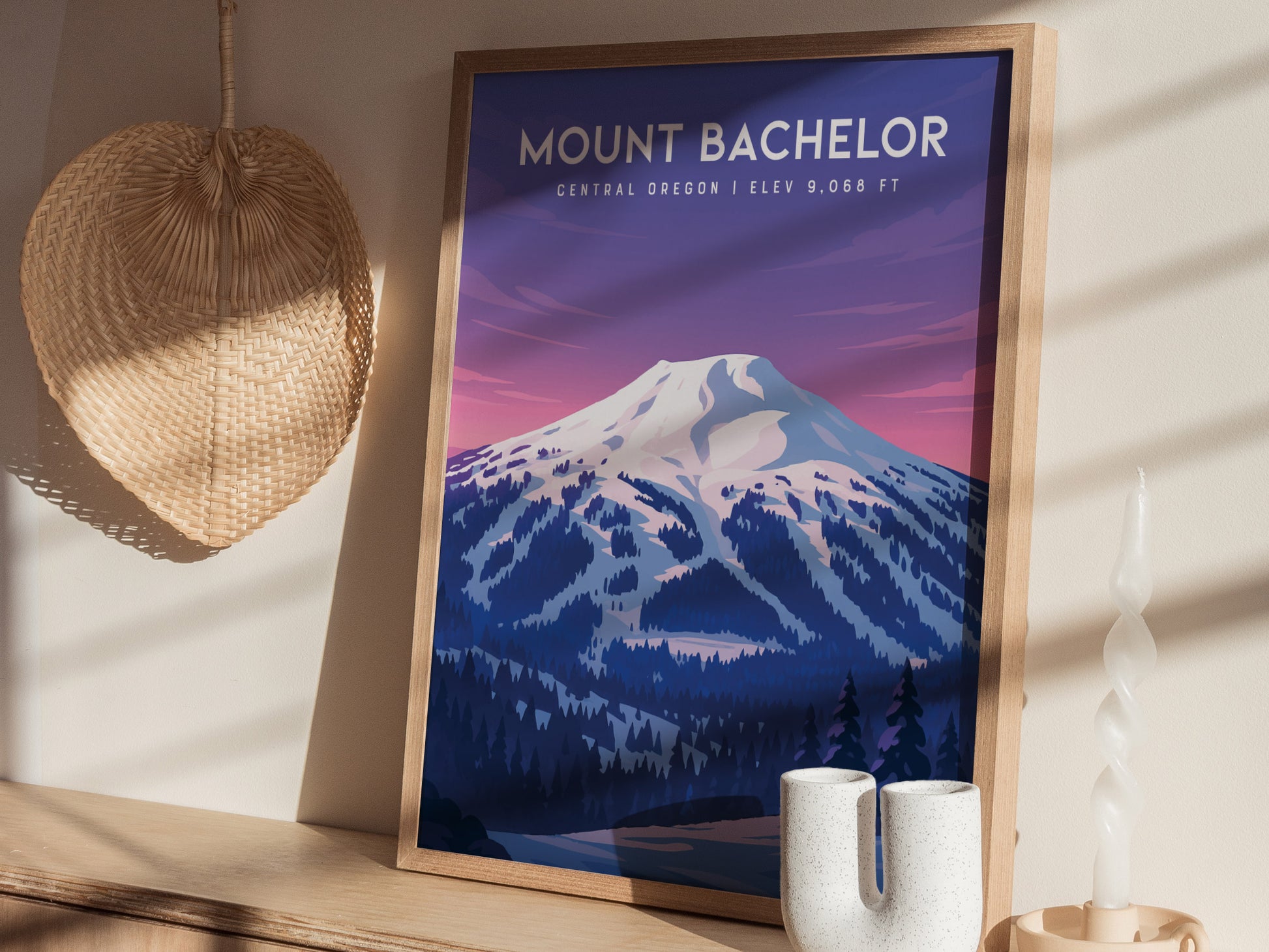 A framed art print of Mount Bachelor in Central Oregon, featuring a stylized depiction of the snow-capped mountain against a twilight sky. The print is displayed on a wooden surface beside decorative vases and a woven wall hanging.