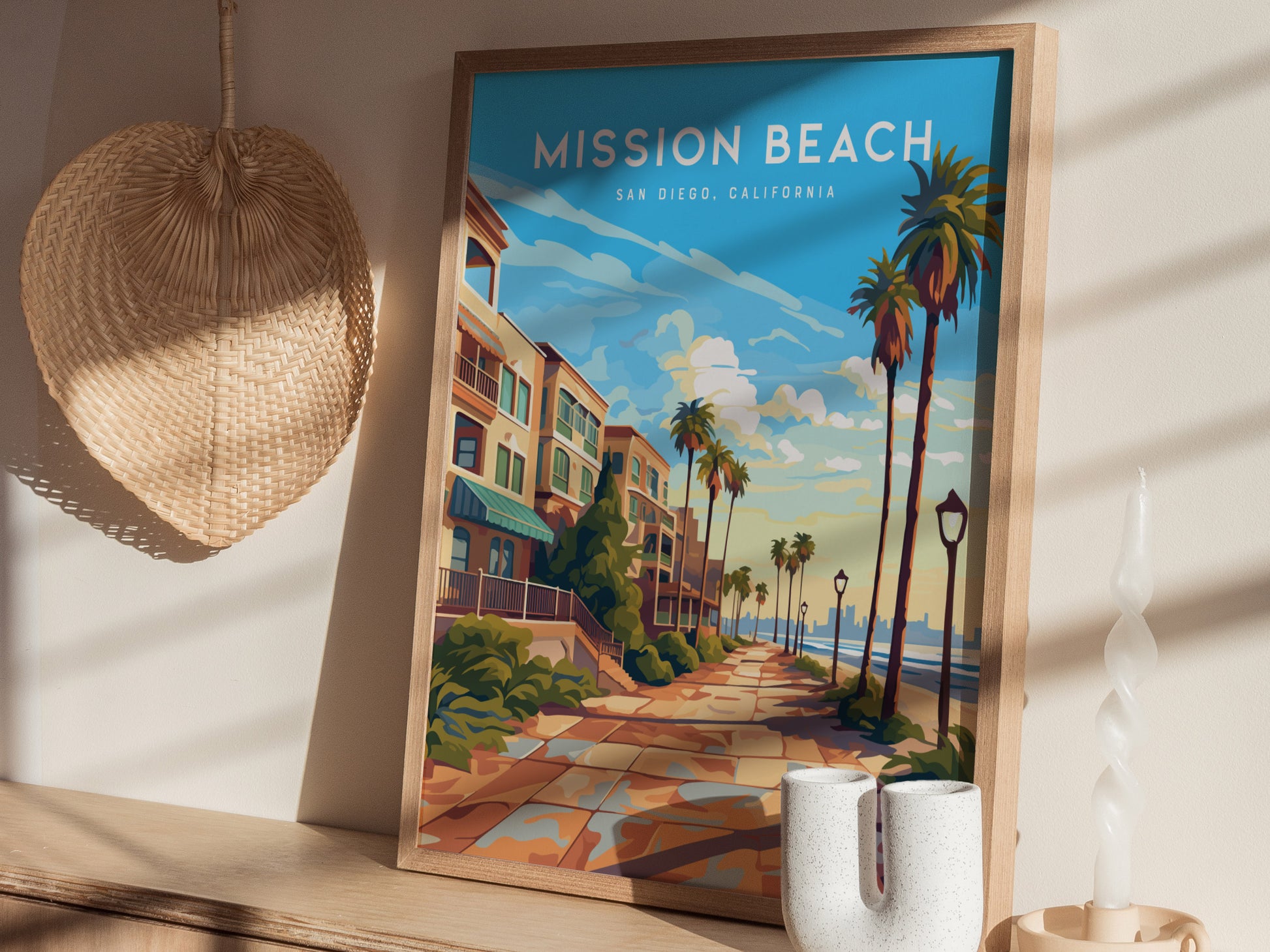 A framed Mission Beach San Diego poster featuring a vibrant boardwalk scene with palm trees is displayed on a wooden shelf, next to a woven fan and decorative candle.