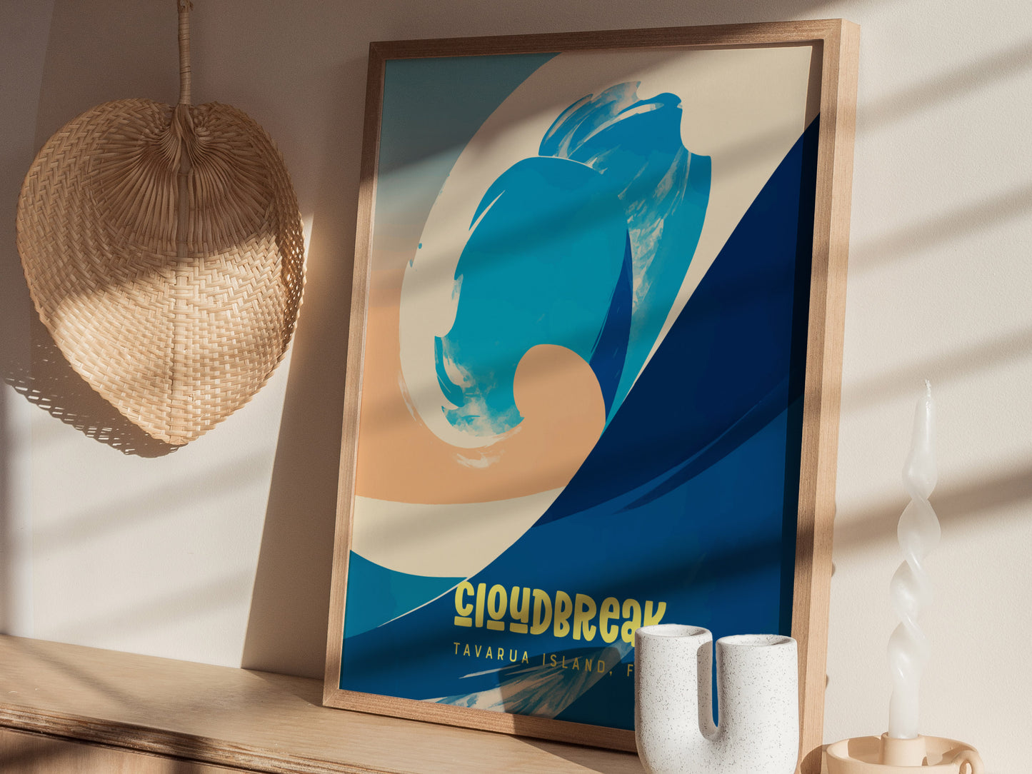 A framed surf poster with abstract blue and beige wave design, titled Cloudbreak Tavarua Island Fiji, is displayed on a wooden surface next to a woven fan and decorative candle.